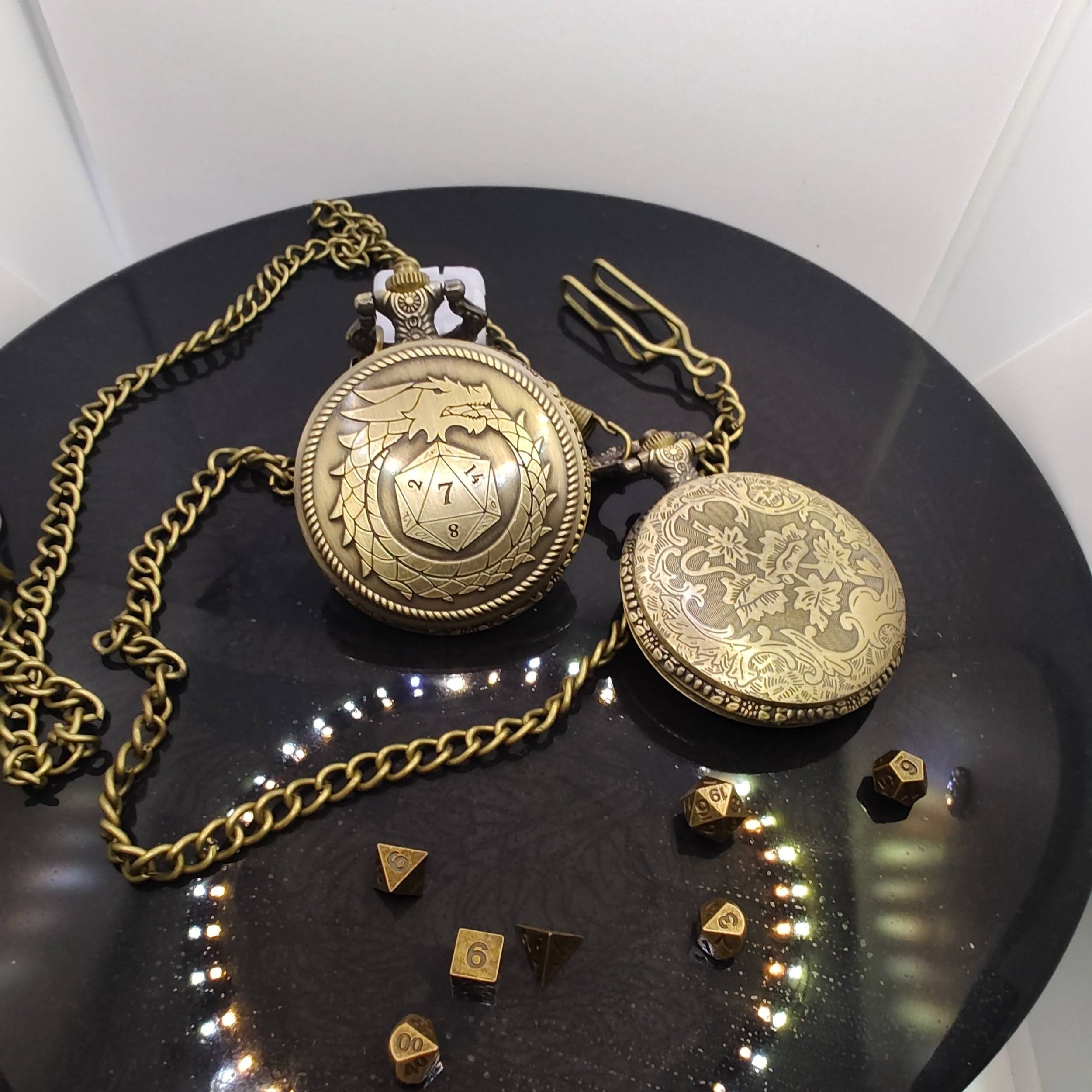 A bronze pocket watch with a dragon motif surrounding a D20 design and a decorative chain, resting on a black surface next to miniature metal dice for DnD and Pathfinder