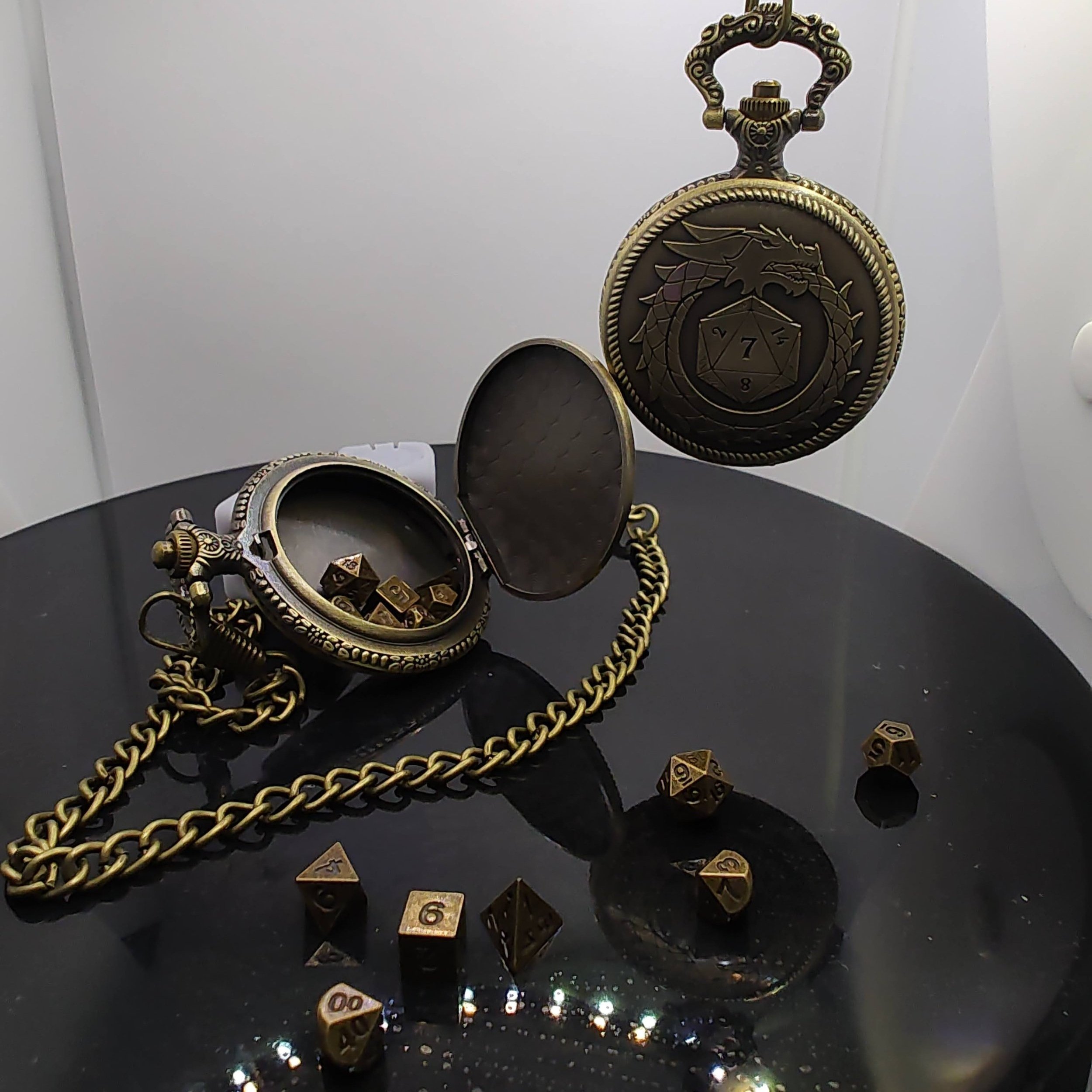 One bronze pocket watch opened to reveal tiny metal dice in a hidden compartment, while another, complete with a chain, showcases a dragon-and-D20 engraving on a black backdrop