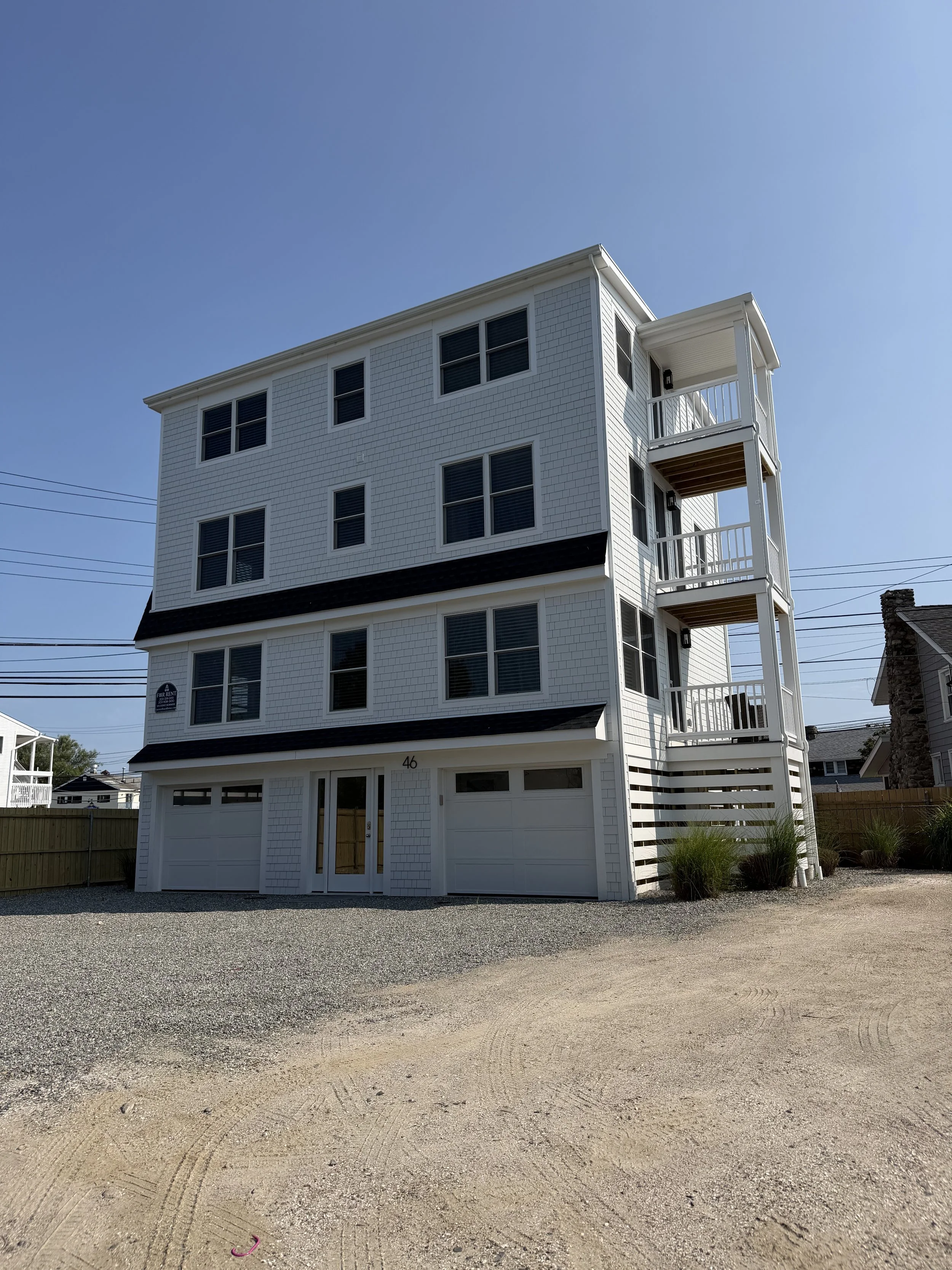 A white, multi-story residential building with a garage on the ground floor and balconies on the upper floors. The building has multiple windows and is set against a clear blue sky. There is a gravel area in the foreground and a wooden fence surrounding the property.
