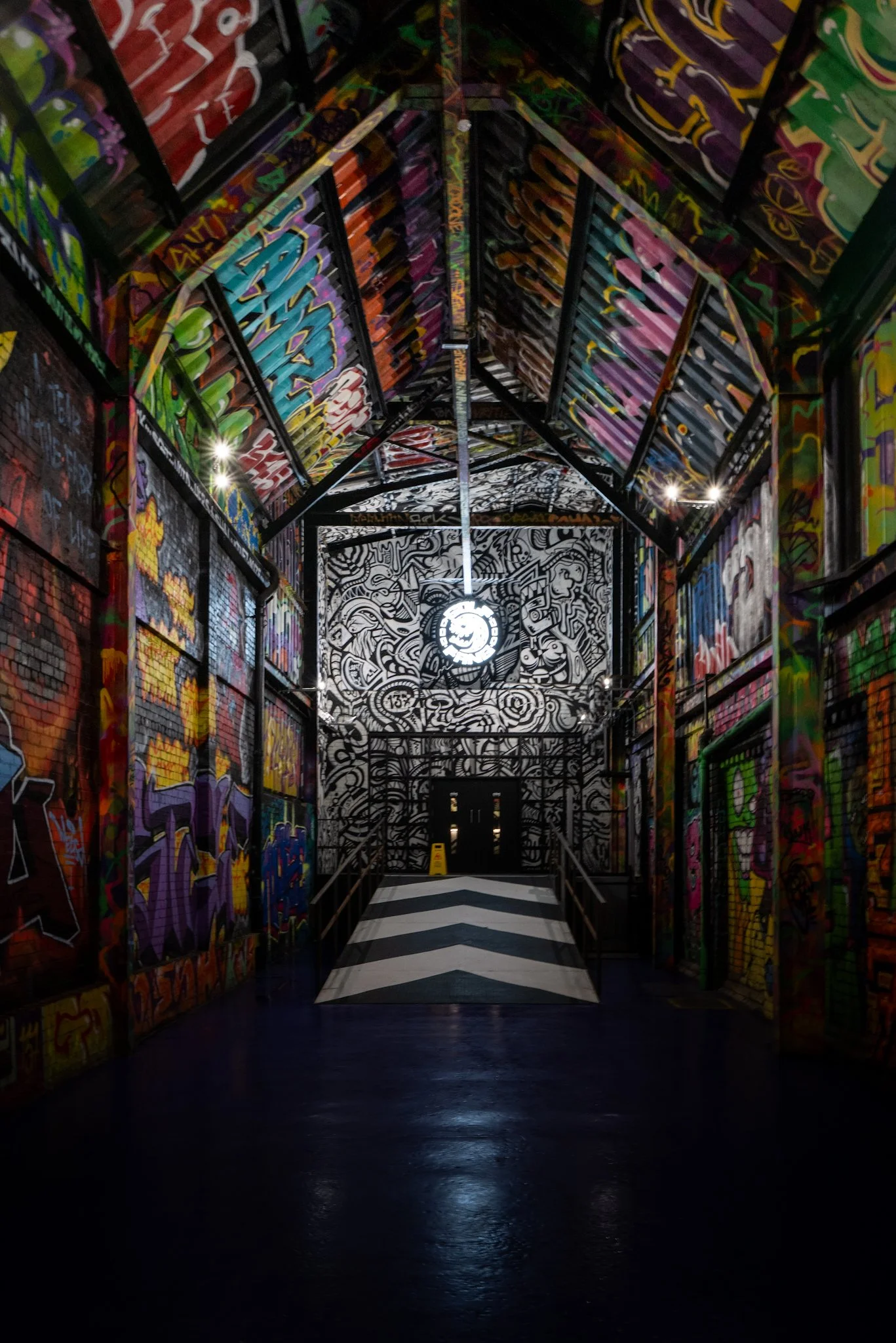 A graffiti-covered indoor space with a black and white striped walkway leading to a double door, illuminated by a central circular light fixture on a high, graffiti-adorned ceiling.