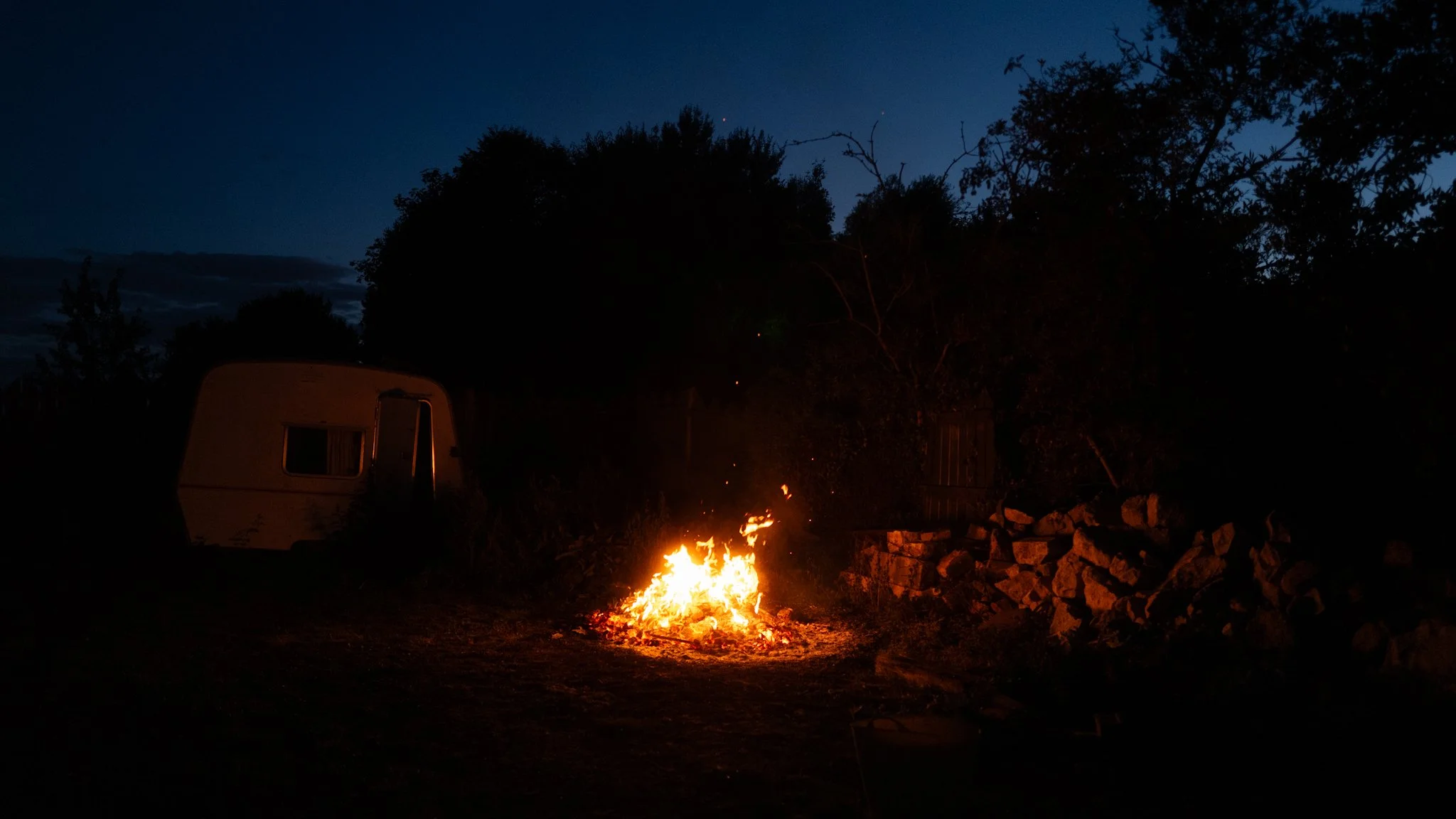 Nighttime outdoor scene with a campfire burning on the ground, surrounded by trees, and an RV trailer parked nearby. The sky is dark with some clouds visible.