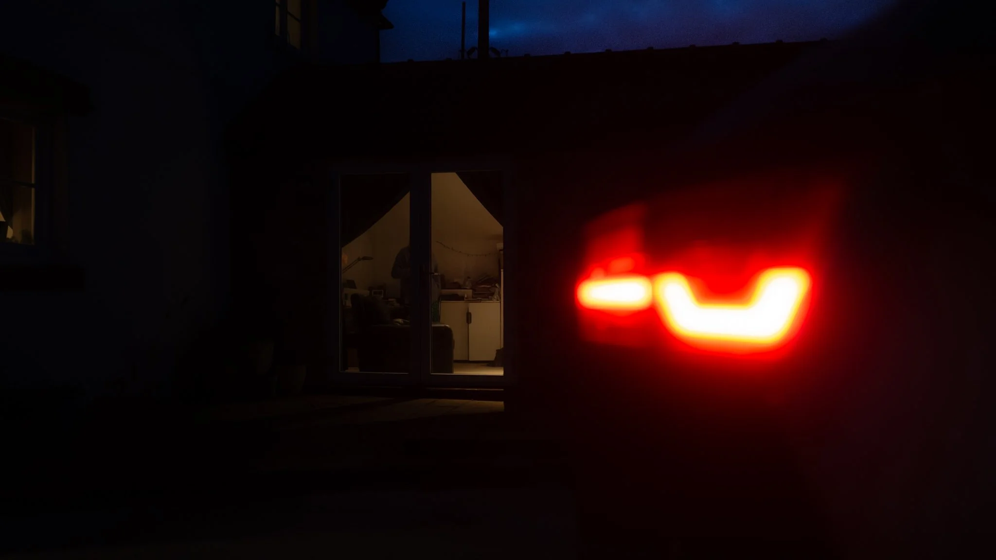 View of a dimly lit house exterior at night with an illuminated alley light and a brightly lit tail light of a vehicle parked outside, with a glass door revealing the interior of the house.