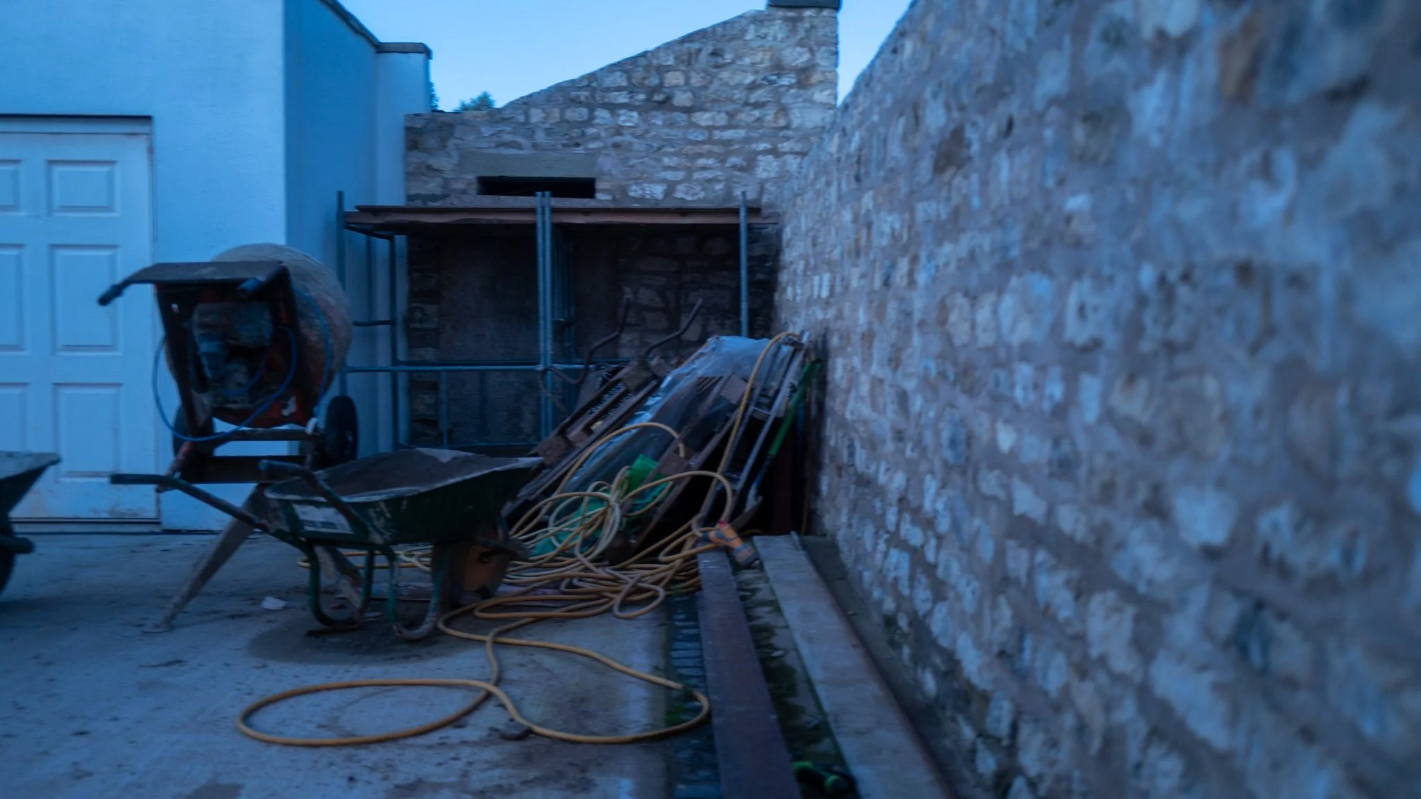 Construction site with cinder block wall, scaffolding, wheelbarrow, and coiled hoses in a backyard.