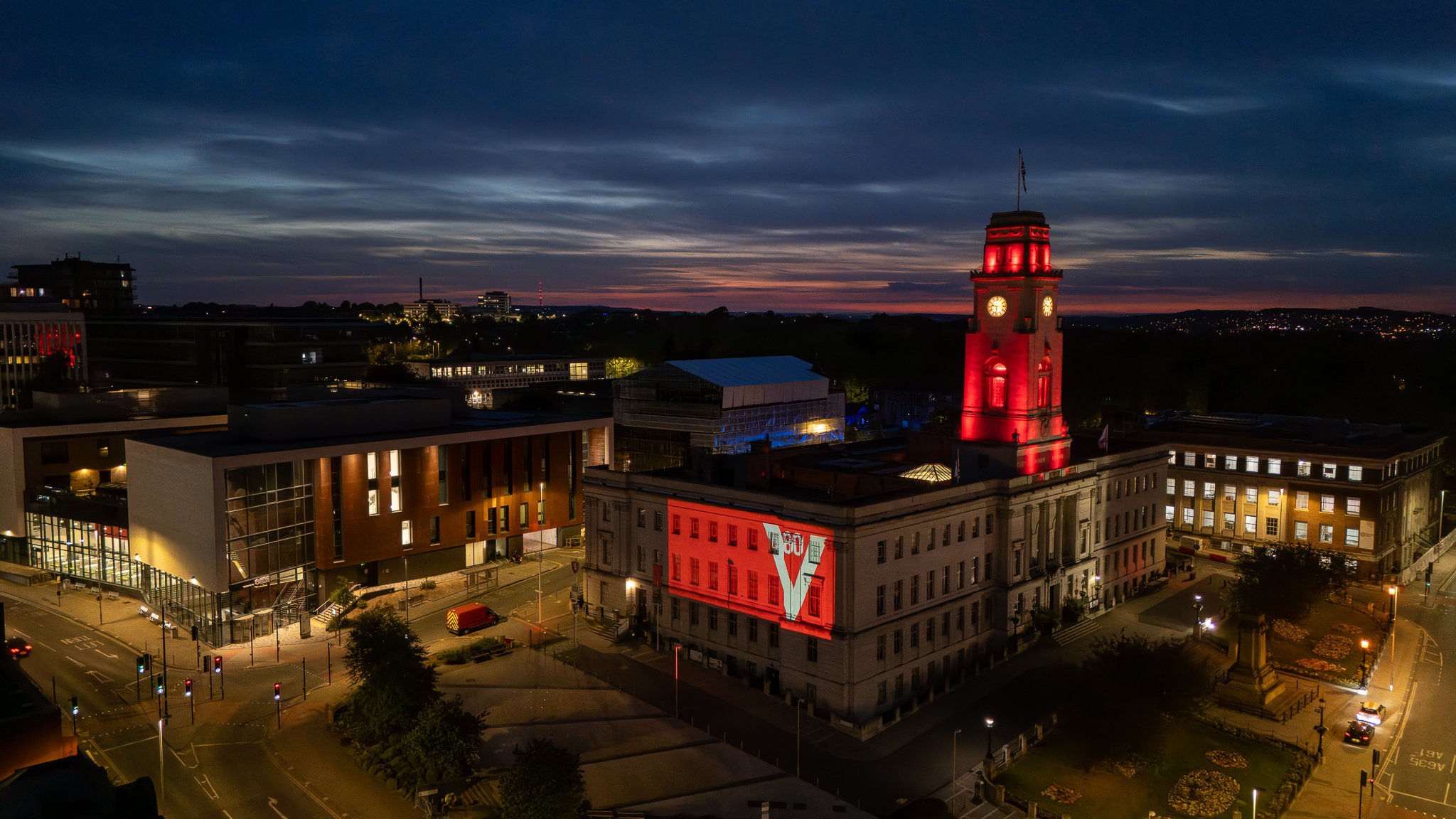 Night view of a city square with illuminated buildings and streets, featuring a prominent historic building with a clock tower brightly lit in red and projective illuminated with the word "LOVE" and a large letter "V" on its facade.