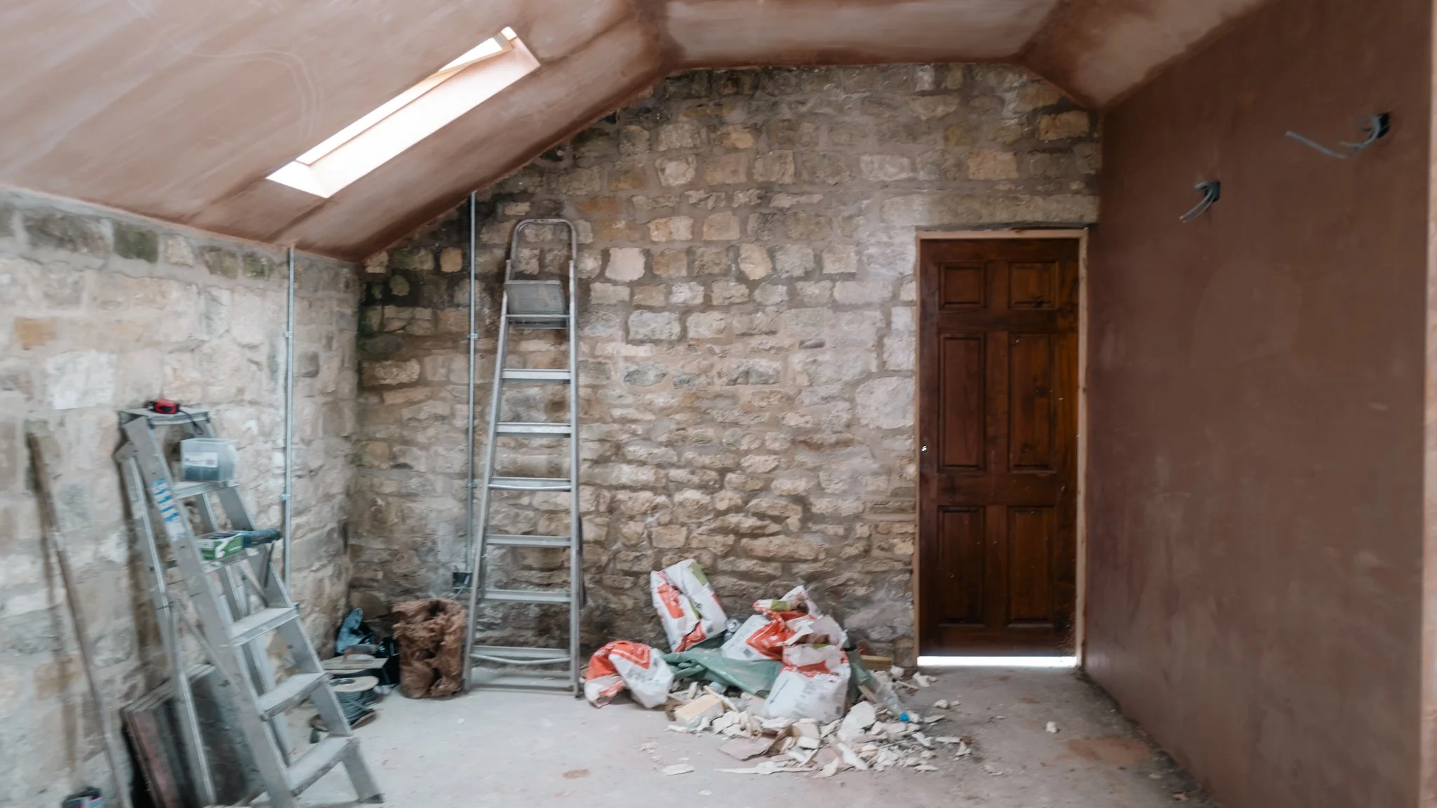An attic room under renovation with unfinished stone walls, a sloped ceiling with a skylight, a closed wooden door, a metal ladder, construction materials, and debris on the floor.