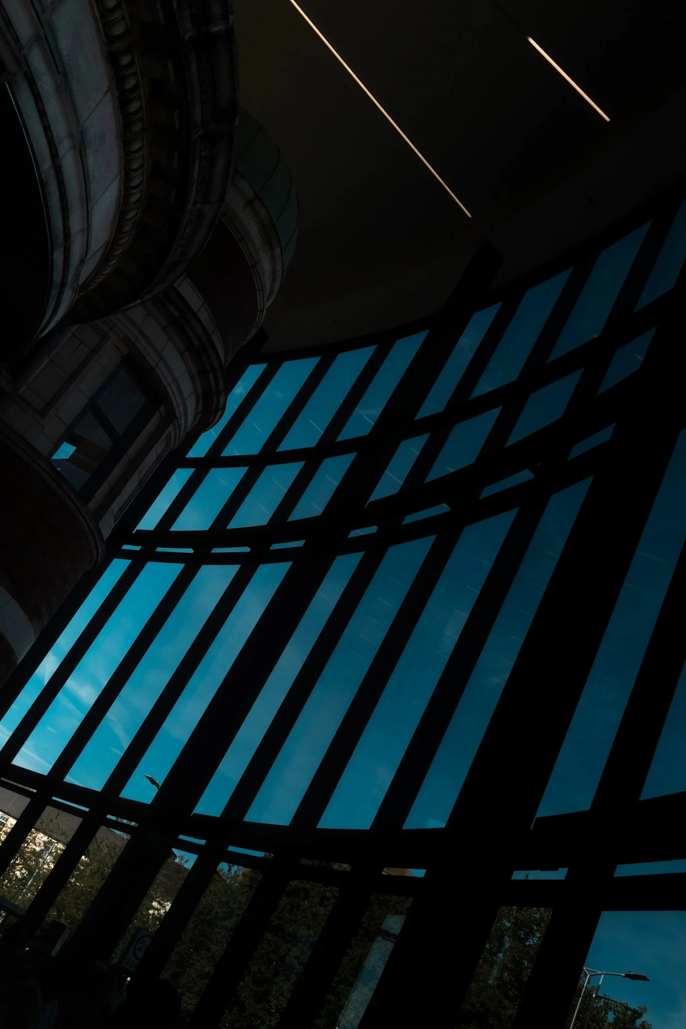Interior view of a modern building with large, angled glass windows and a curved balcony, showing blue sky and clouds outside.