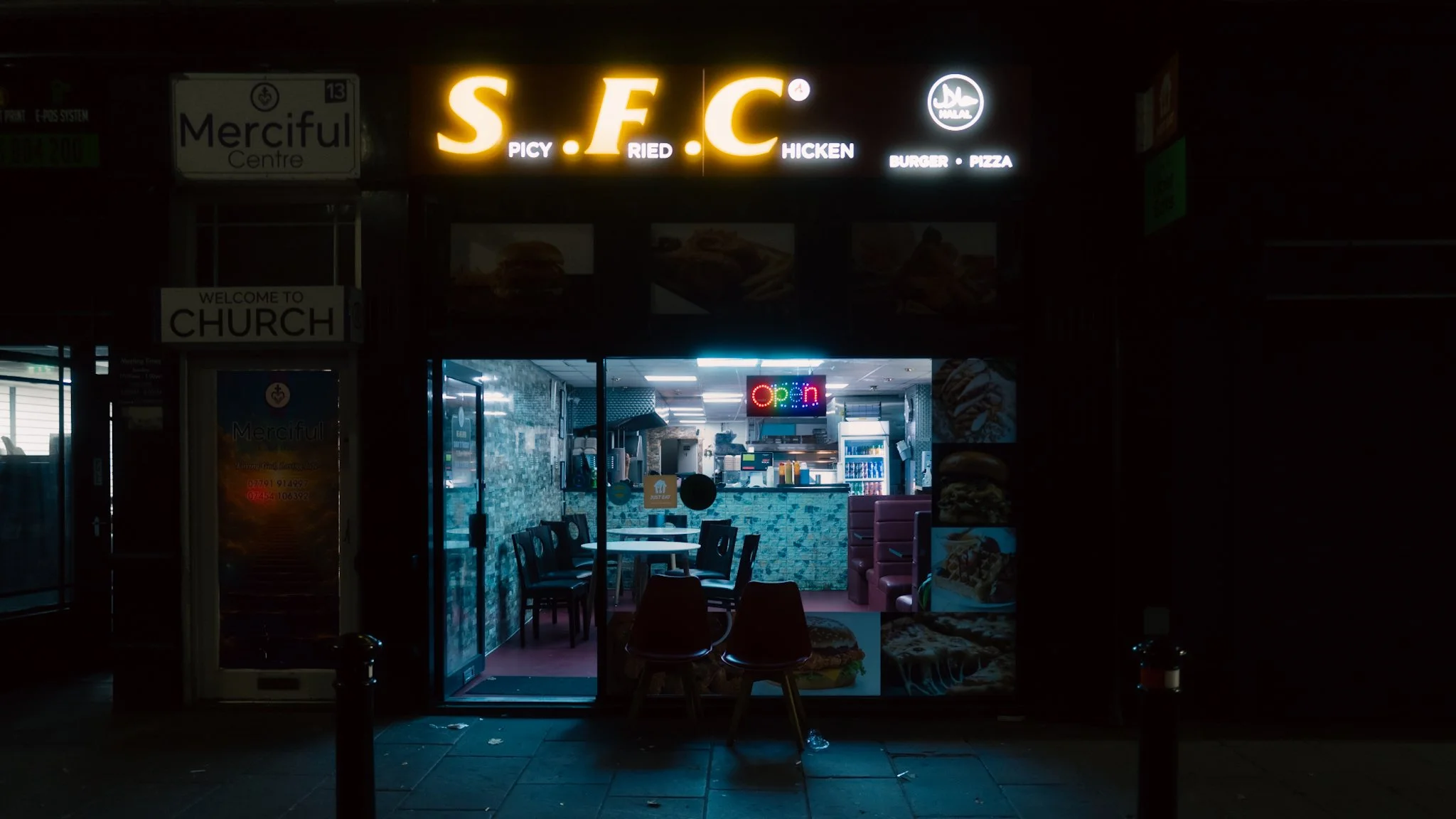 Small fast food restaurant with illuminated sign displaying 'S.F.C' and descriptions like 'Spicy Fried Chicken' and 'Burger Pizza'. The interior is visible through the glass door, with an 'Open' neon sign and a cozy seating area with chairs and table