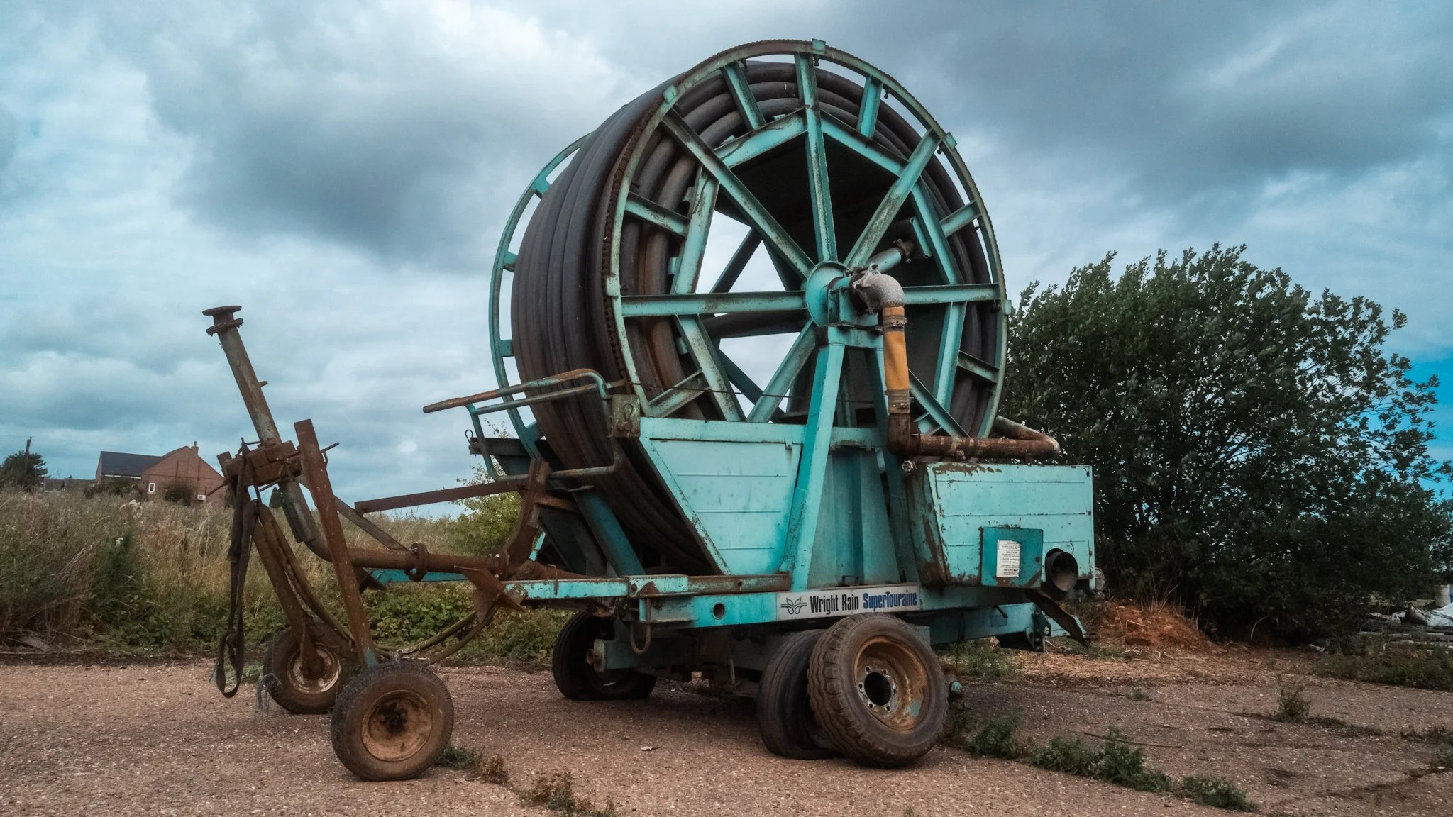 An old, rusty irrigation reel machine with a blue frame and large wheel, parked outdoors on a dirt surface near bushes and trees.
