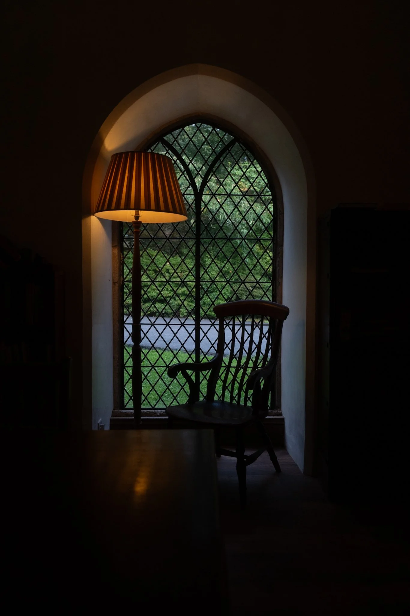 A dimly lit room with a wooden chair and a floor lamp beside an arched window with a diamond lattice pattern, overlooking greenery outside.