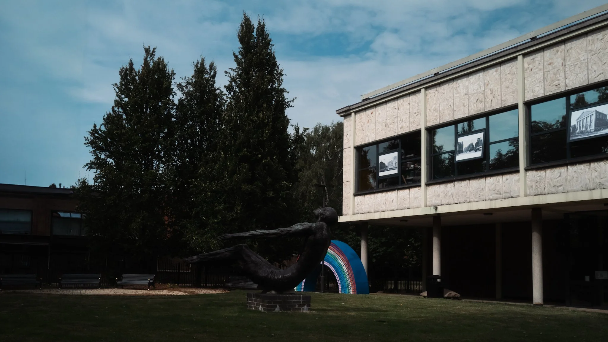 A bronze sculpture of a person diving, located in front of a modern building with large windows and architectural sketches in the windows. Two trees are visible behind the sculpture and building, with a partly cloudy sky above.