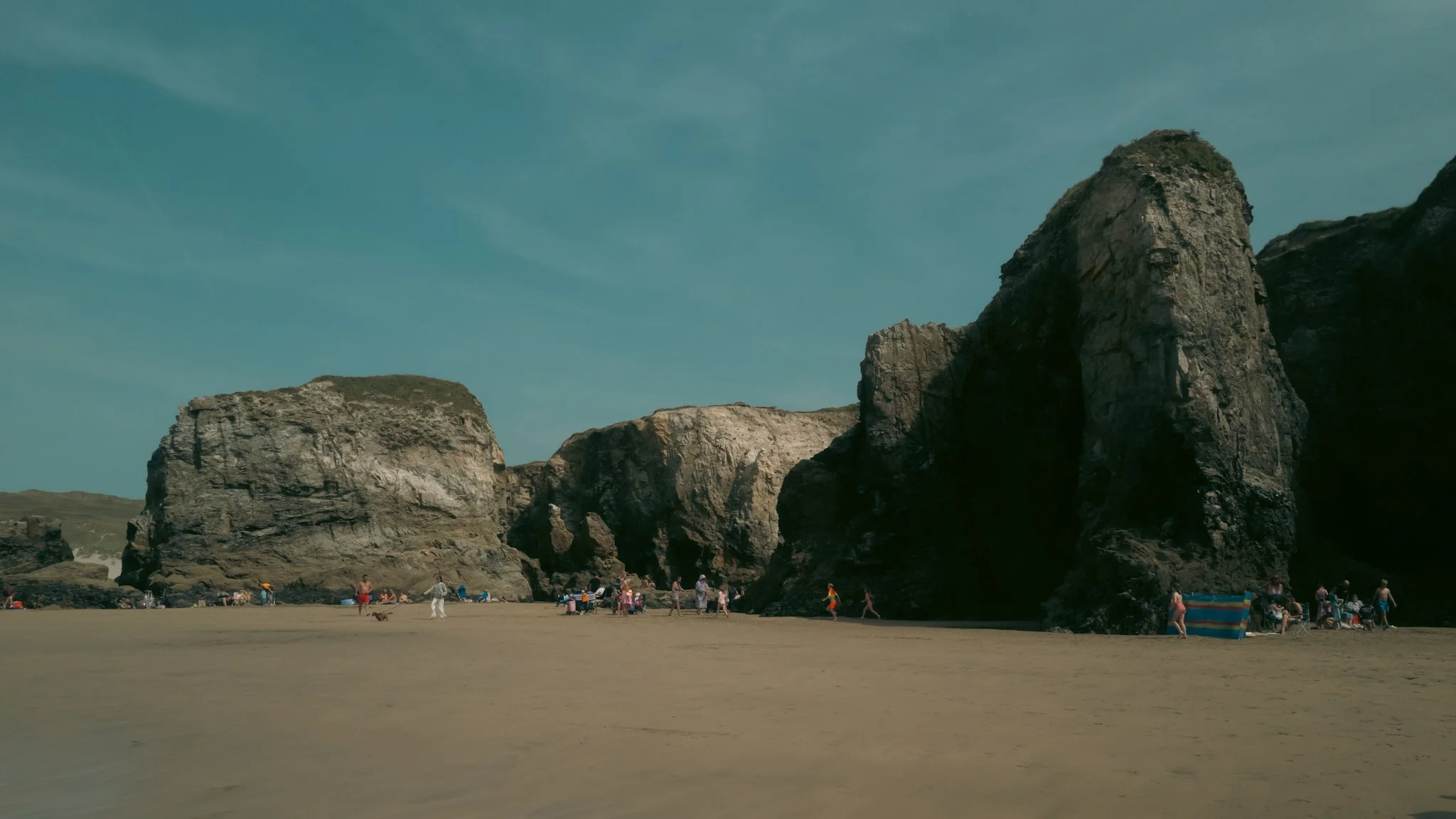 Beach with large rock formations, people relaxing and walking on the sand, clear sky.