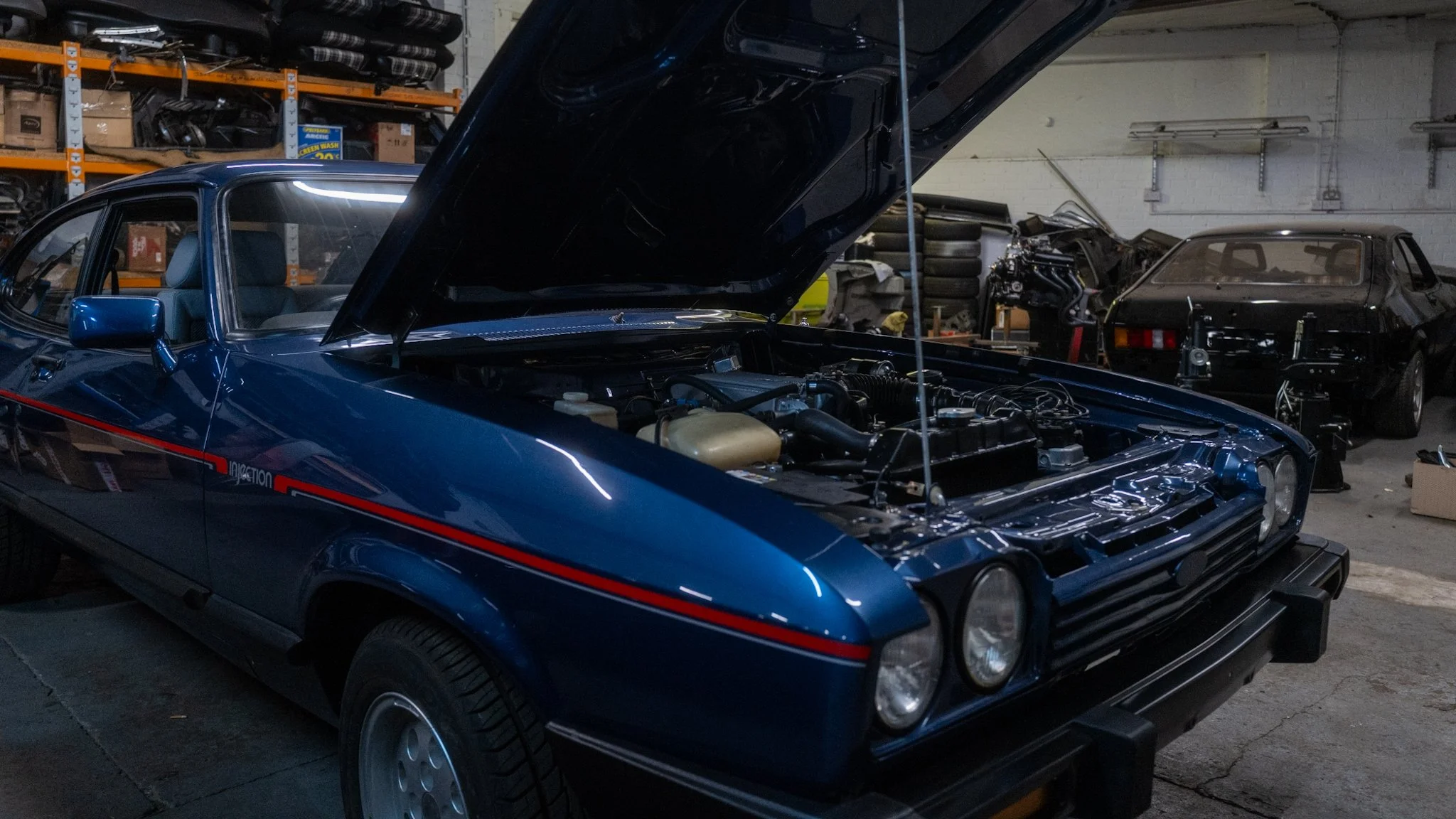 A blue classic car with its hood raised inside a garage or workshop, with other vehicles and various car parts in the background.