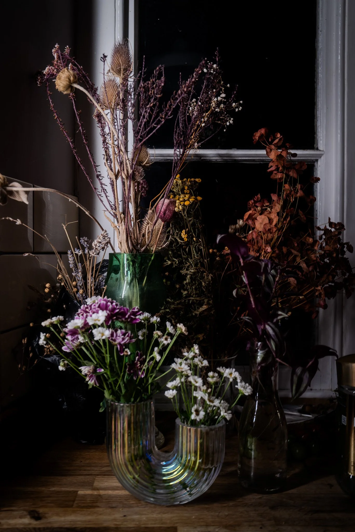 Vases with dried and fresh flowers on a wooden surface in front of a window at night.