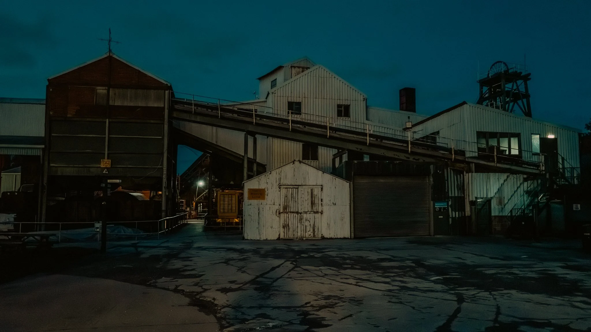 Old industrial building at night with metal structures and a small white shed in front, faint lighting and dark sky.