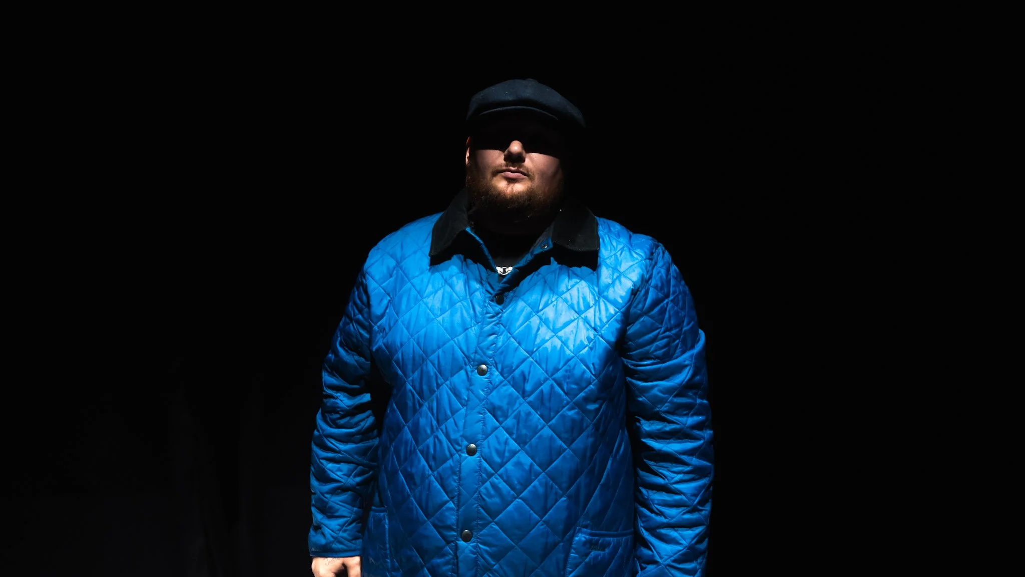 A man wearing a blue quilted jacket, black hat, and black shirt stands in front of a black background with dramatic lighting.