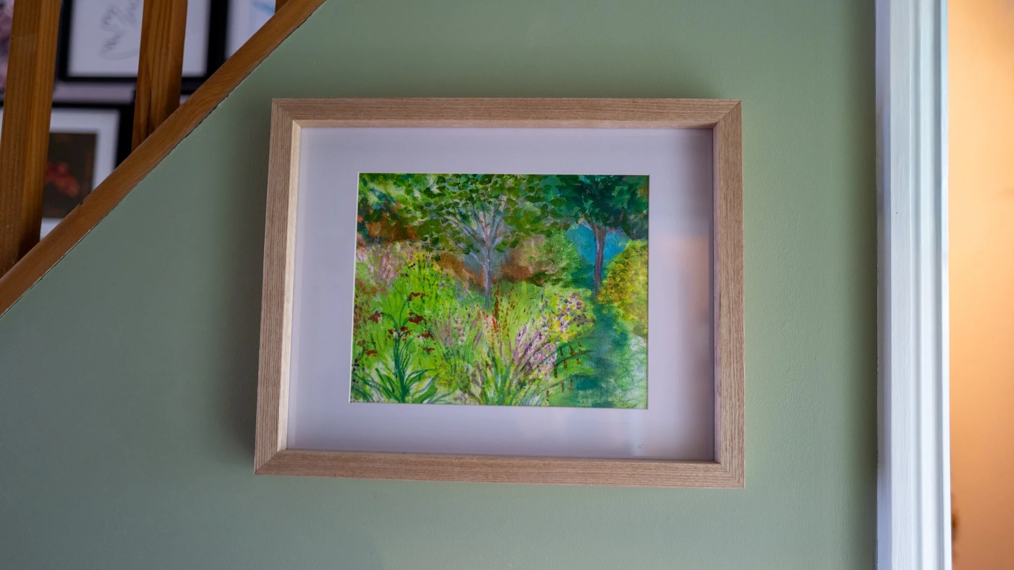 Colorful watercolor painting of a lush garden scene with trees, greenery, and flowers, framed in light wood and hung on a green wall near a staircase.