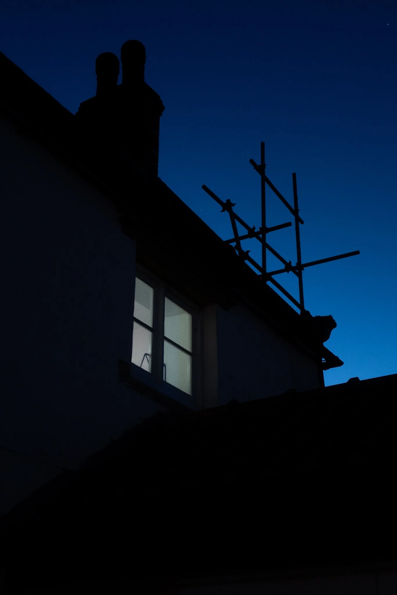 The silhouette of a house’s roof with two chimneys and a window visible, against a blue twilight sky. There is also an image of scaffolding on the roof.