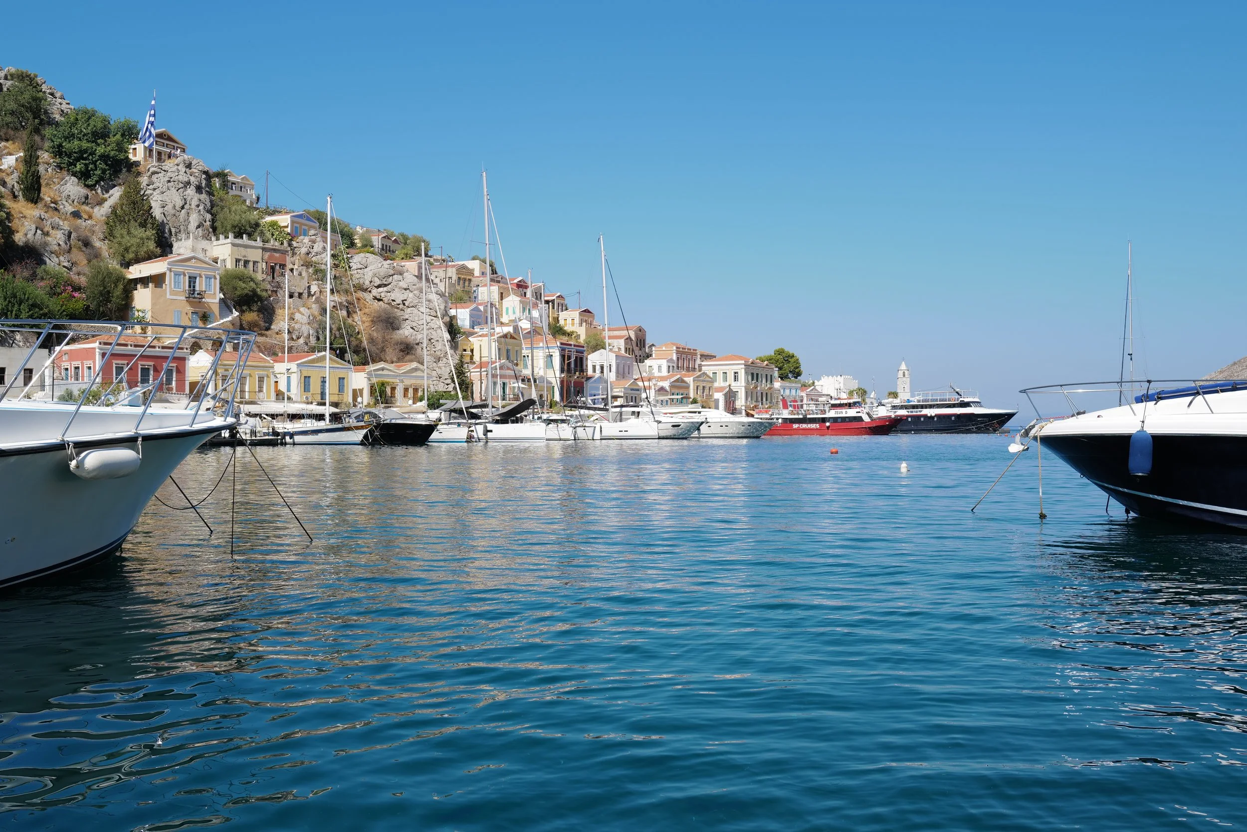 View of a harbor with sailboats and yachts docked, with colorful houses and buildings on a hillside in the background under a clear blue sky.