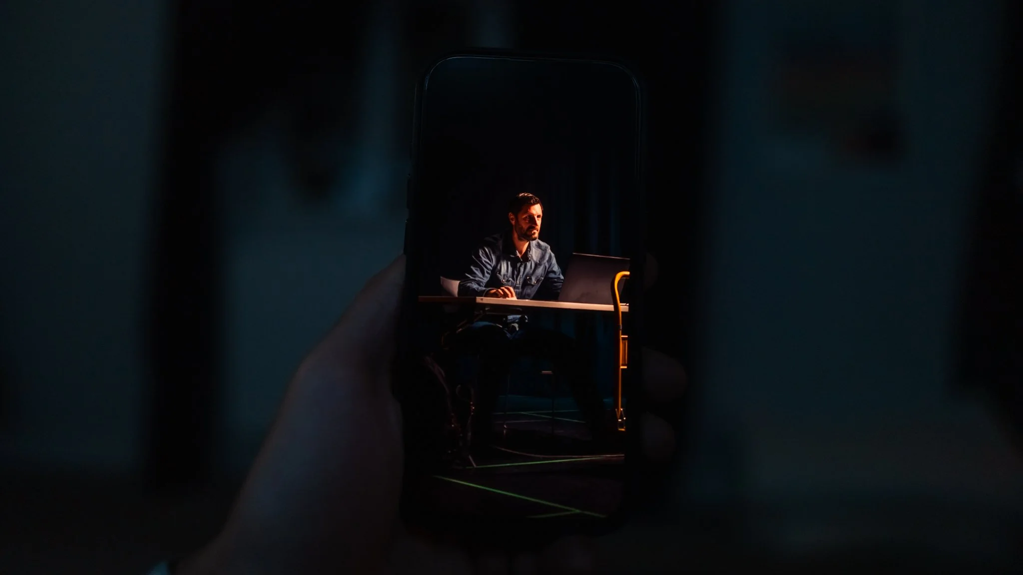 Person sitting at a desk, using a laptop, seen through the reflection on a smartphone screen in a dark environment.