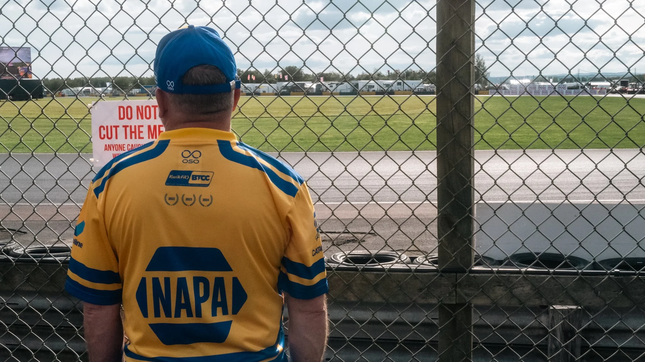 A person wearing a yellow and blue INAPAM shirt and a blue cap standing behind a chain-link fence at a racetrack, watching the event. There is a sign on the fence that says "Do not cut the mesh."