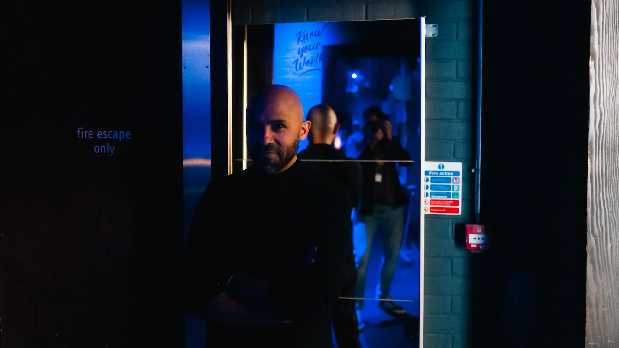 A bearded man in dark clothing standing in front of a doorway with blue lighting. Reflection in the mirror behind him shows other people and a sign that says 'Know Your Way.' To the left, a sign says 'fire escape only.'