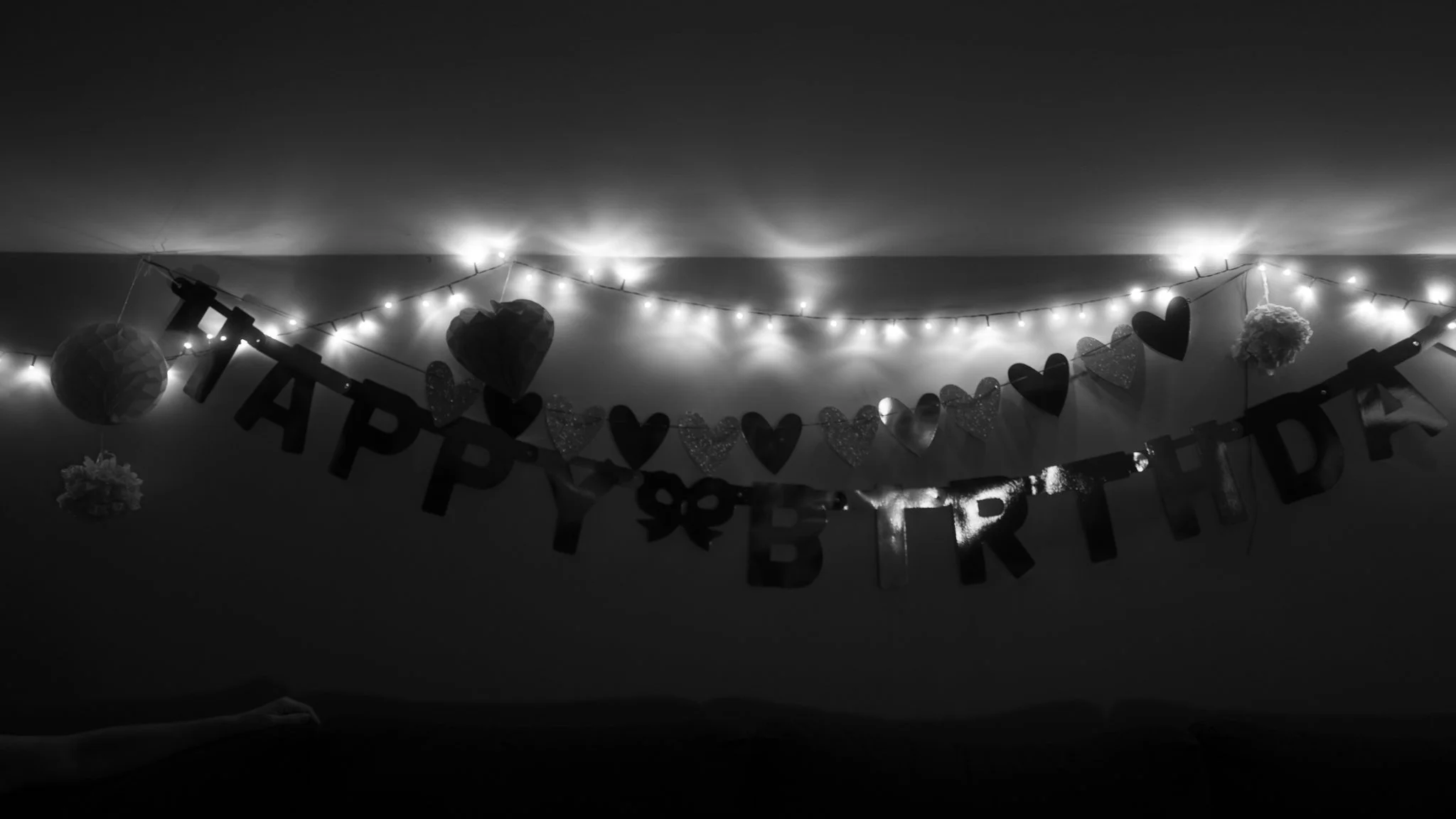 Black and white photo of a birthday celebration with string lights, heart-shaped decorations, and a 'Happy Birthday' banner on the wall.