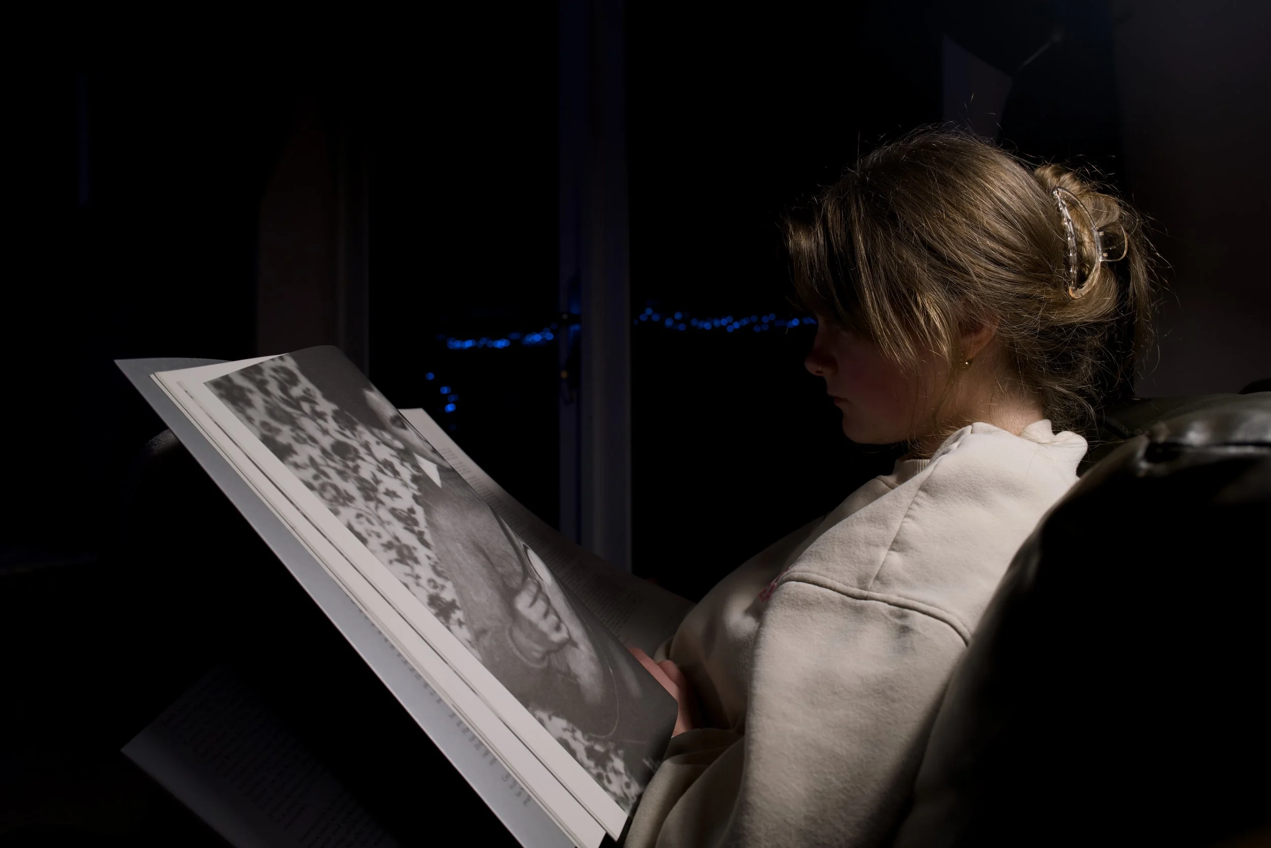 A young girl reading a magazine with black-and-white photos, sitting in a dimly lit room at night.