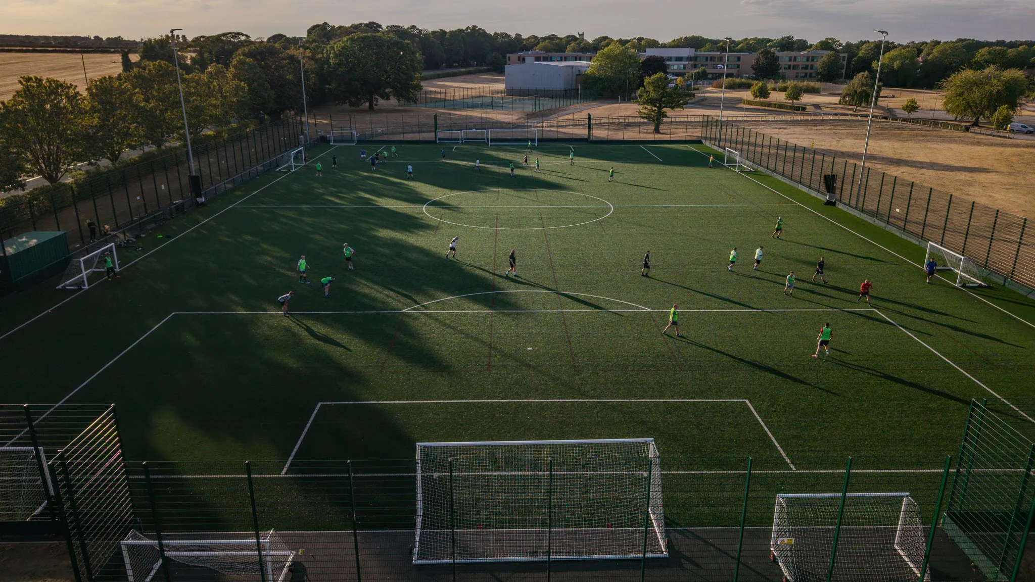 An aerial view of a soccer field with players practicing or playing a game during late afternoon or early evening. The field is surrounded by a tall fence and has goalposts at each end. There are trees, a parking lot, and a few buildings in the background.