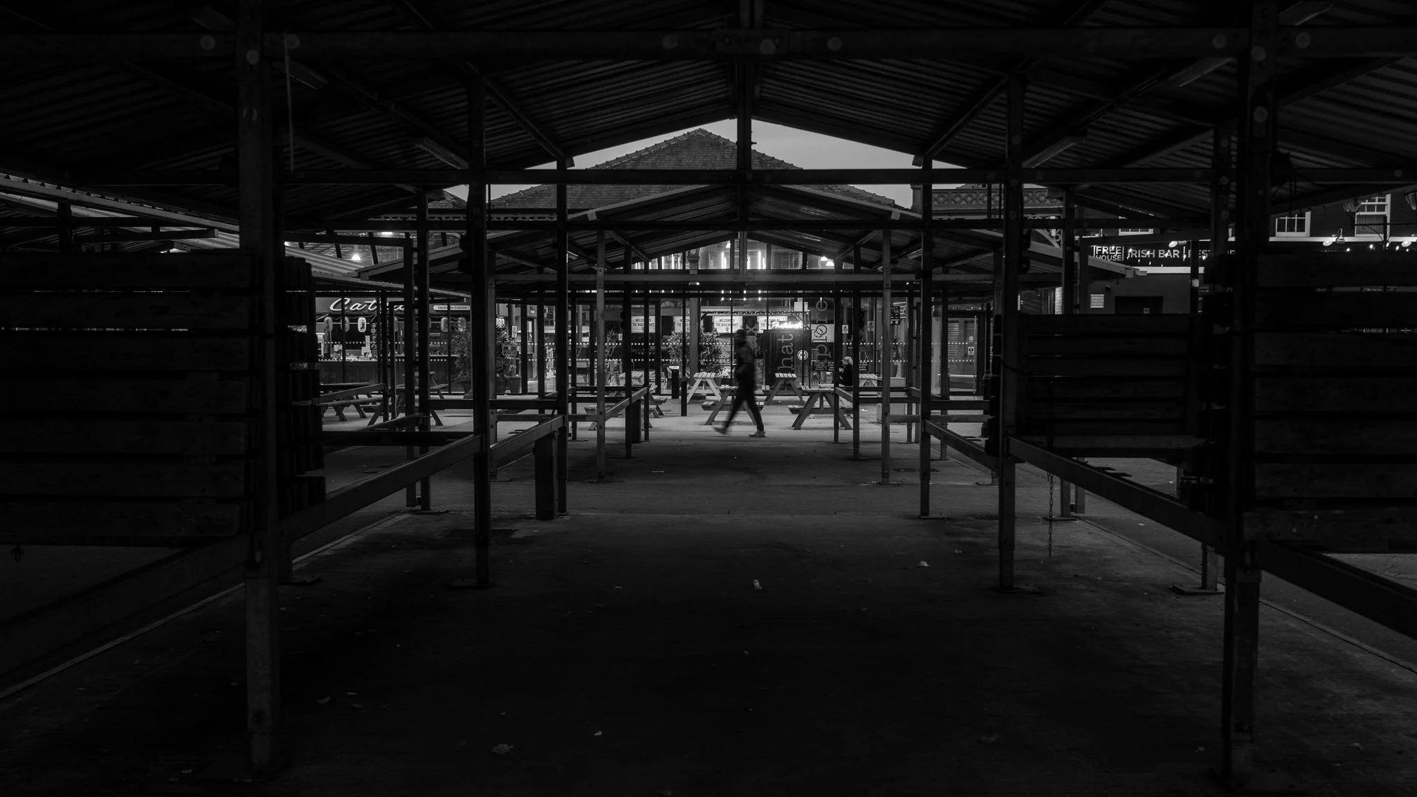 Empty outdoor market with metal scaffolding and a person walking, in black and white.