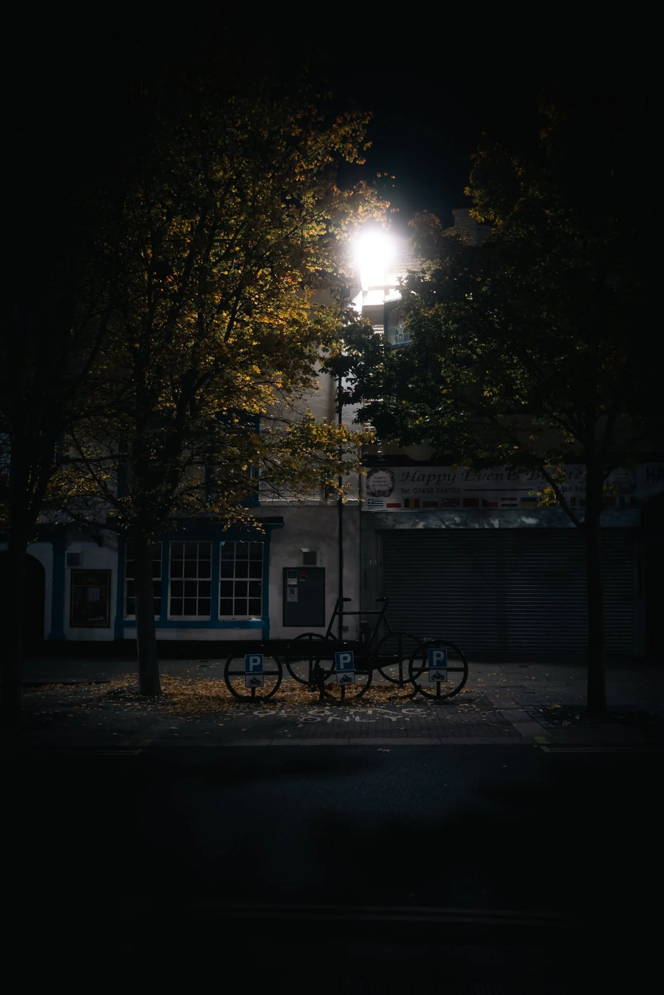 Nighttime street scene with a bright streetlight illuminating trees, a bicycle rack, and storefronts with closed shutters.