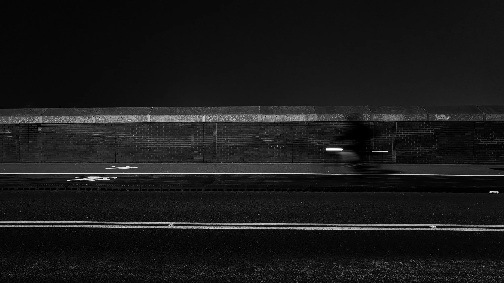 Nighttime street scene with a brick wall and a nearby cyclist in motion, captured in black and white.
