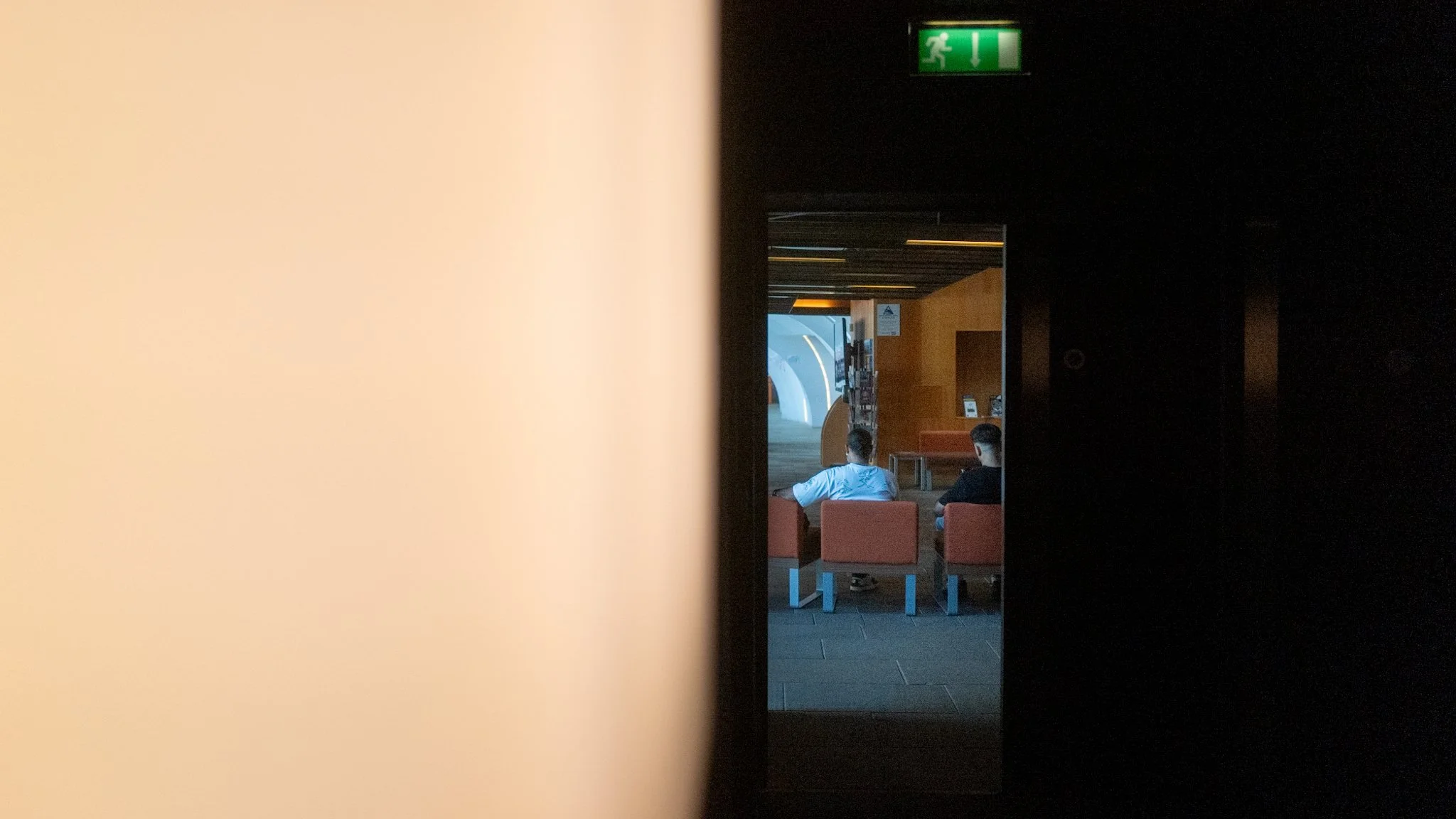 View through a narrow opening showing two people seated on orange chairs in a lobby or waiting area, with a bright blue abstract sculpture and wooden elements in the background.