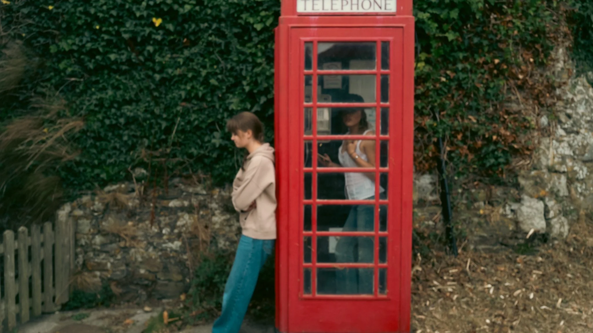 A young man leaning against a traditional red British telephone booth while a woman inside the booth looks at her phone outdoors.