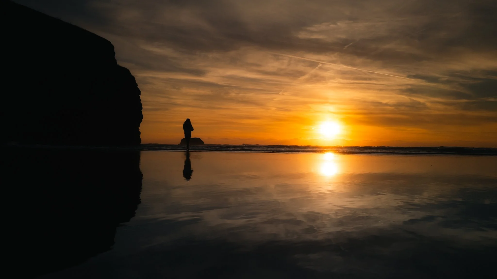 Silhouette of a person standing on the beach at sunset, with a large rock formation on the left and the sun low on the horizon reflecting on the wet sand.