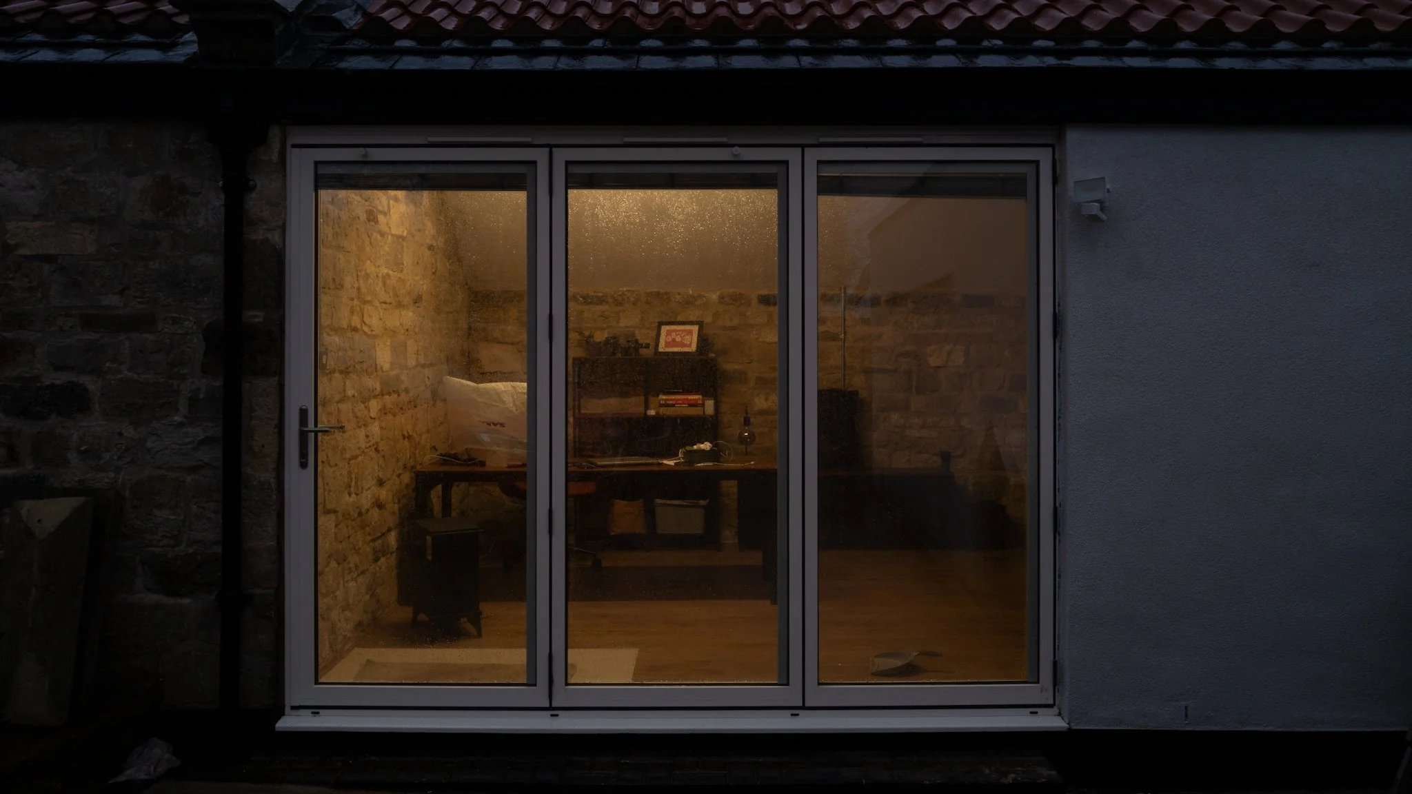 A rear view of a house with a large glass sliding door showing a lit interior room with brick walls and a wooden floor.