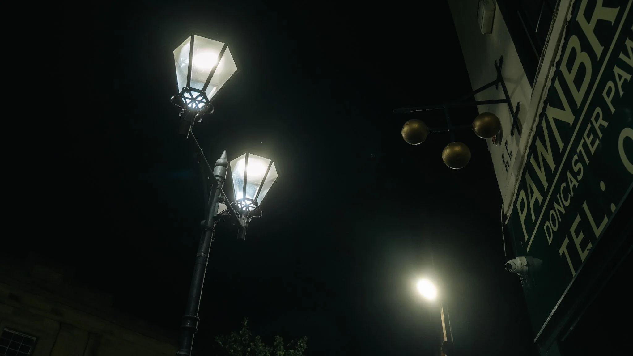 Night scene with two old-fashioned street lamps illuminated, a wall with hanging black spherical lights, and a 'Pianist' poster partially visible on the right.