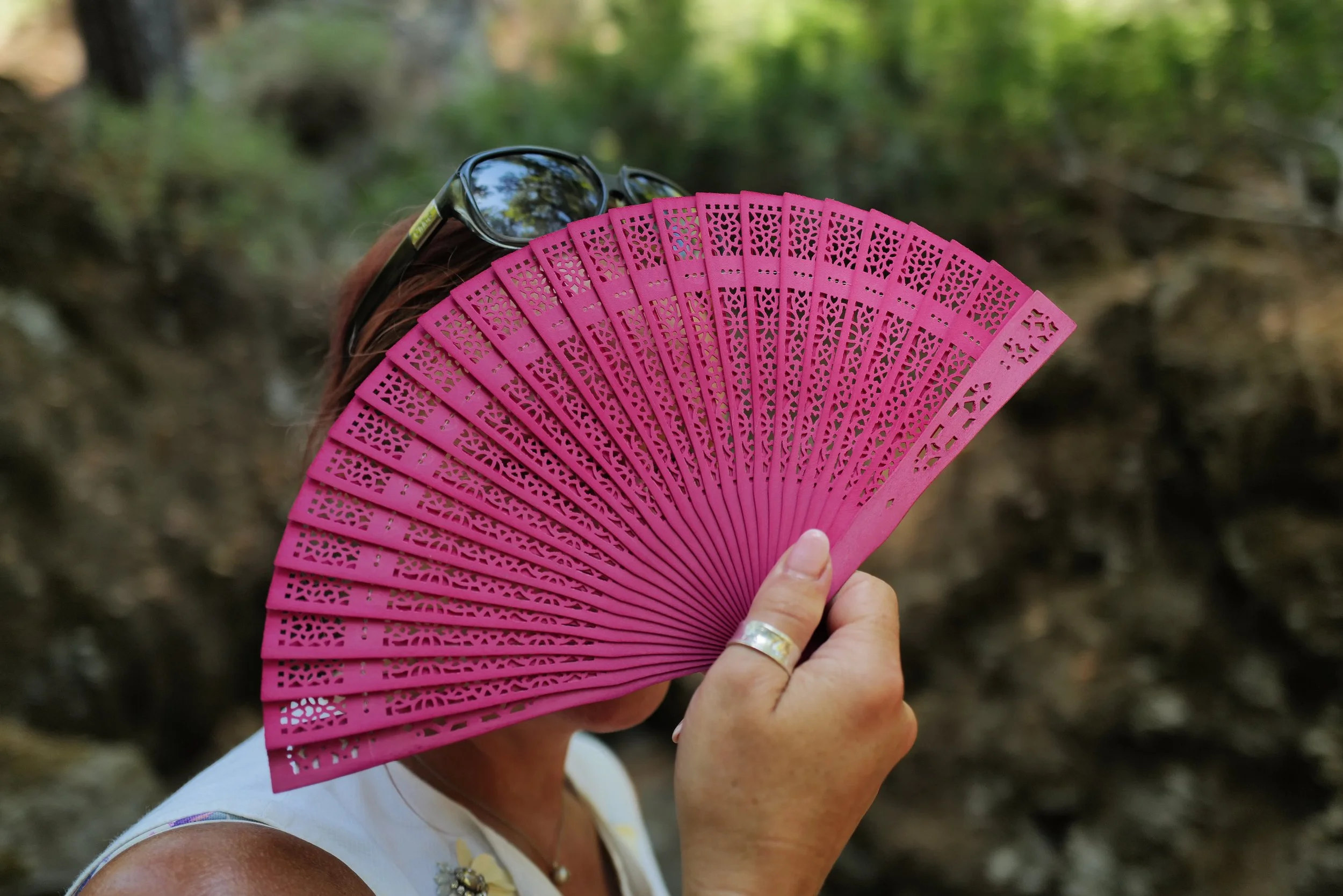 Person holding a pink decorative fan covering their face and sunglasses, outdoors in a natural setting.