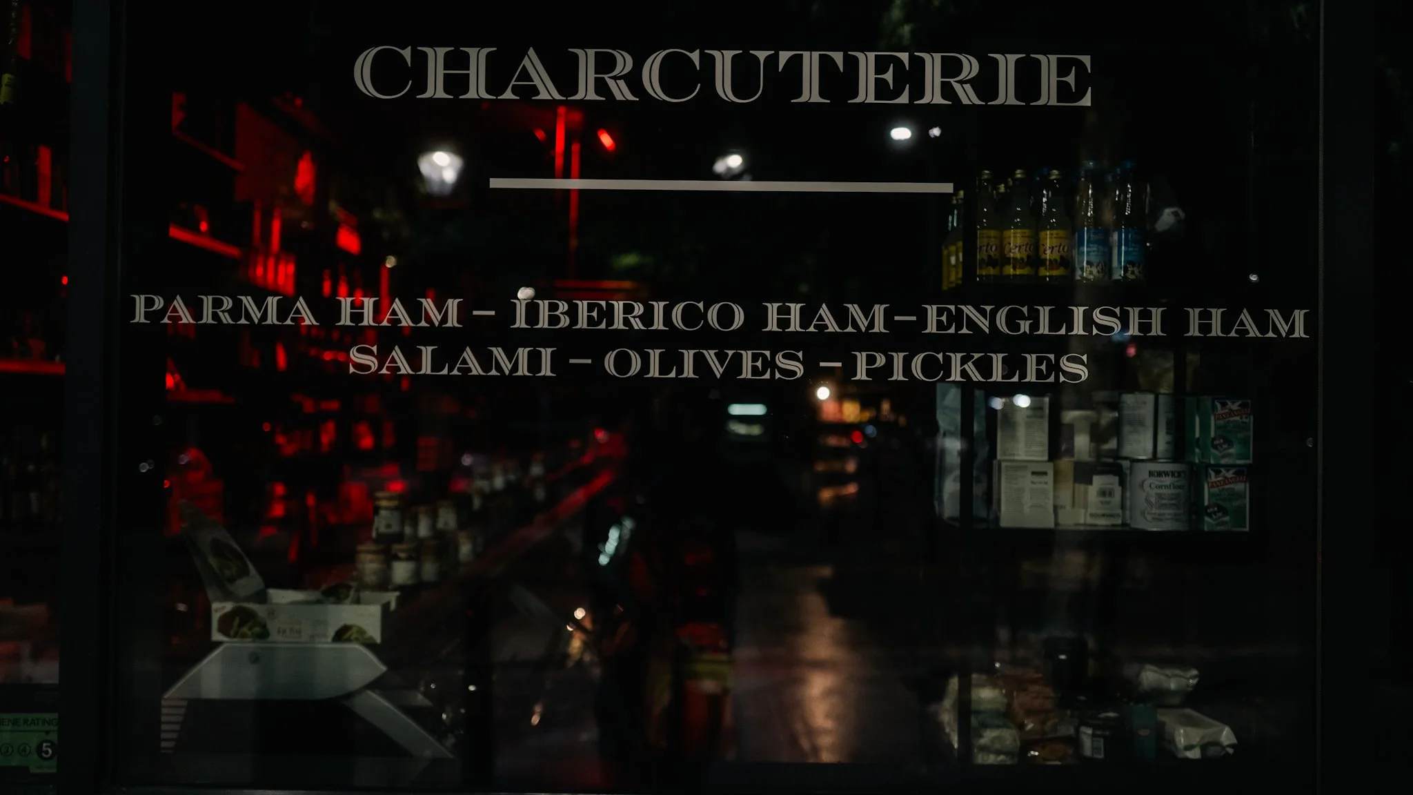 A glass window with a menu for a charcuterie shop, listing items like Parma ham, Iberico ham, English ham, salami, olives, and pickles, with shelves of bottles and packaged goods inside.