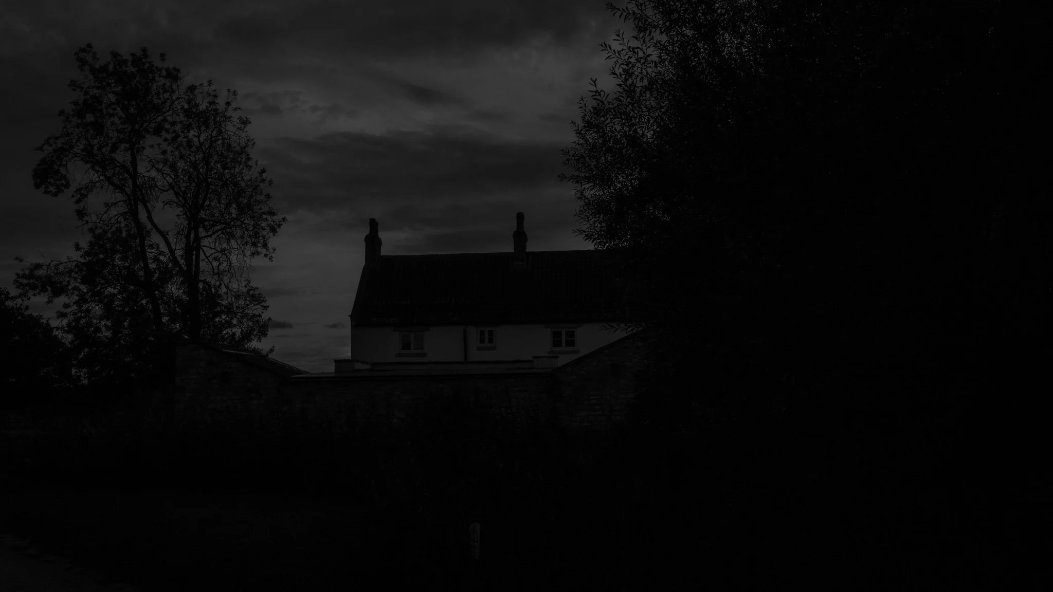 A dark, nighttime image of a house with a sloped roof, surrounded by trees, with a cloudy sky overhead.