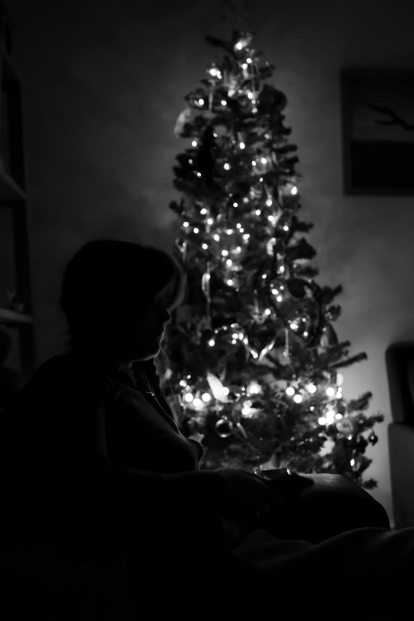 Silhouette of a person sitting in front of a lit Christmas tree in a dark room.