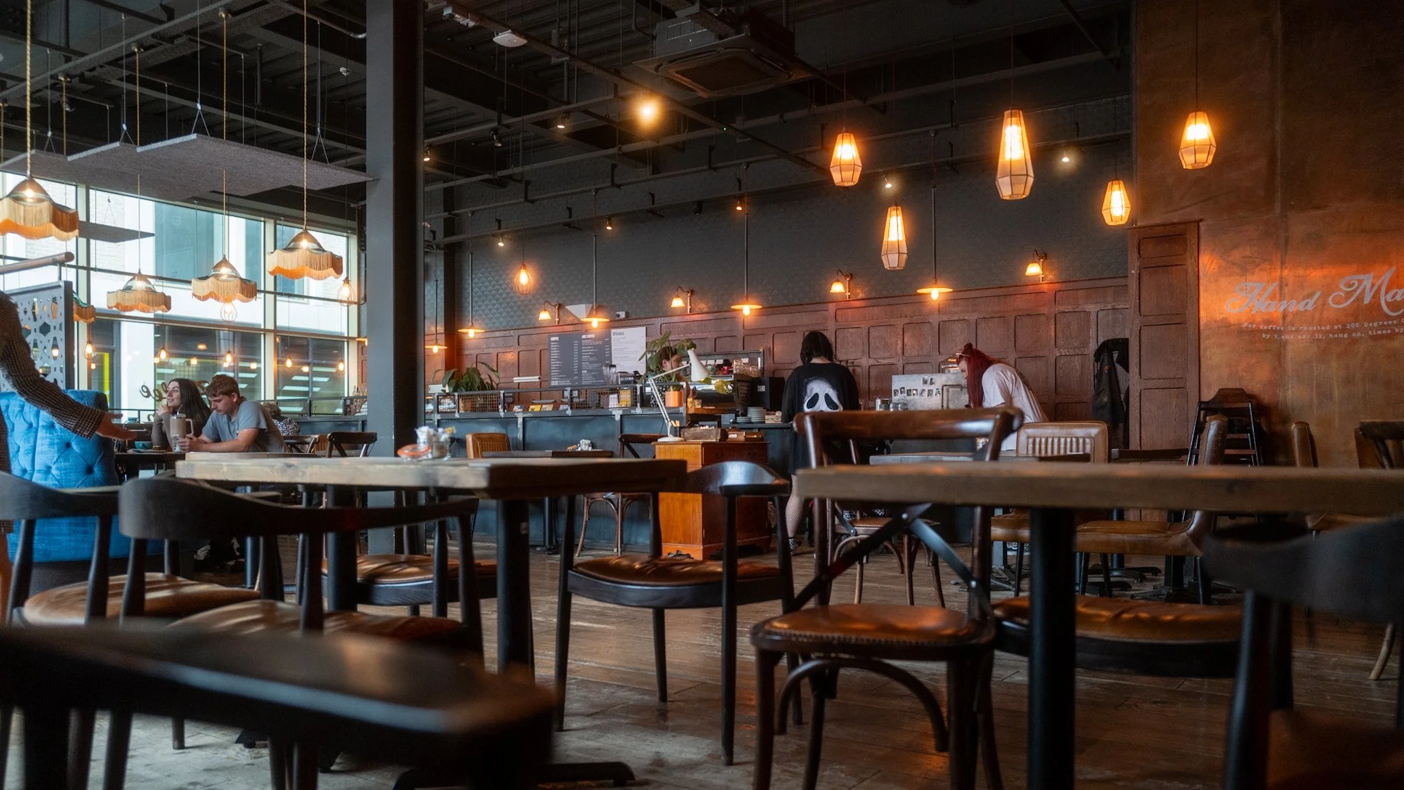 Interior of a cozy cafe with wooden tables and chairs, warm lighting from pendant lamps, a large window on the left, and people working or chatting inside.
