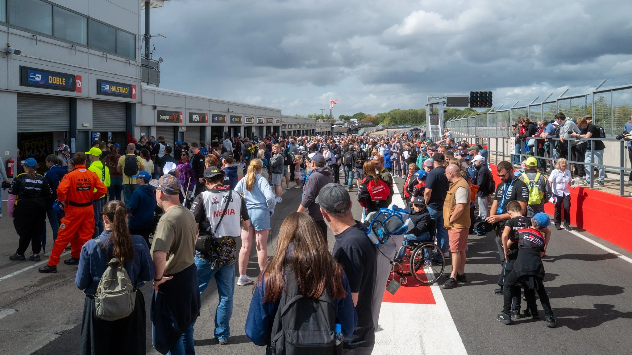 Crowd of people at a motorsport race track paddock area, with teams, fans, and media personnel walking and gathering near garages and pit lane under an overcast sky.