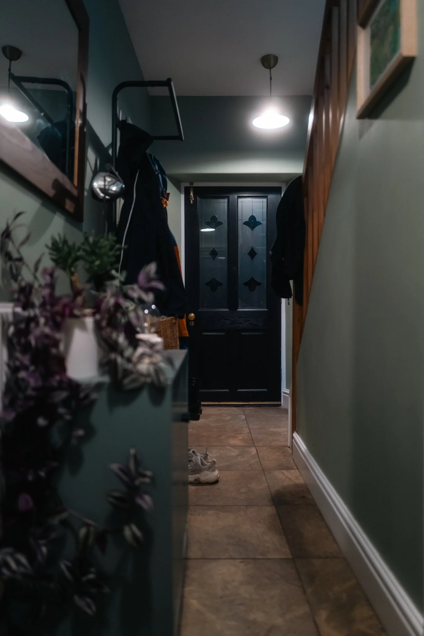 View of a narrow hallway with a black front door at the end, and a pair of sneakers on the tiled floor in front of the door. The hallway has green walls, a wooden staircase on the right, a small dresser with flowers on the left, and coat hooks with c