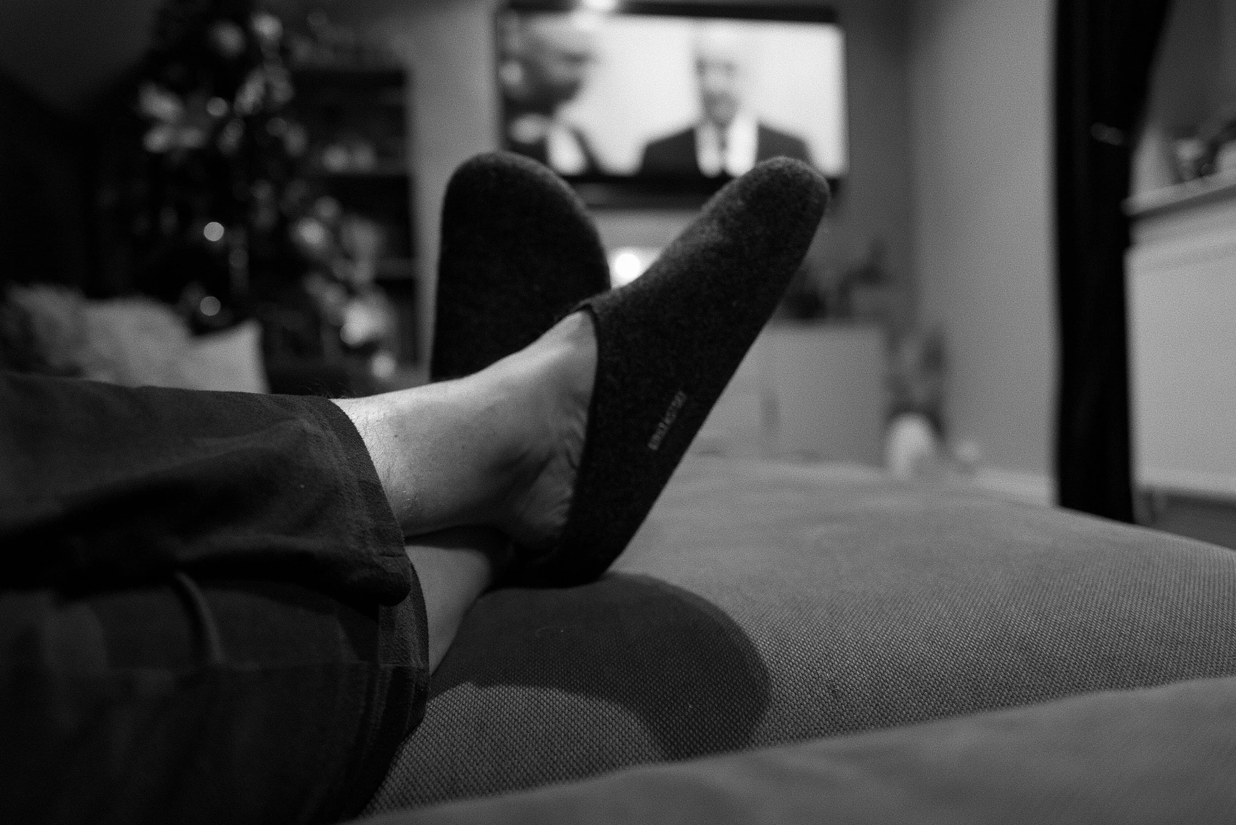 A person lying on a couch with their legs extended, wearing a sock on one foot, in a living room with a television and shelves in the background.