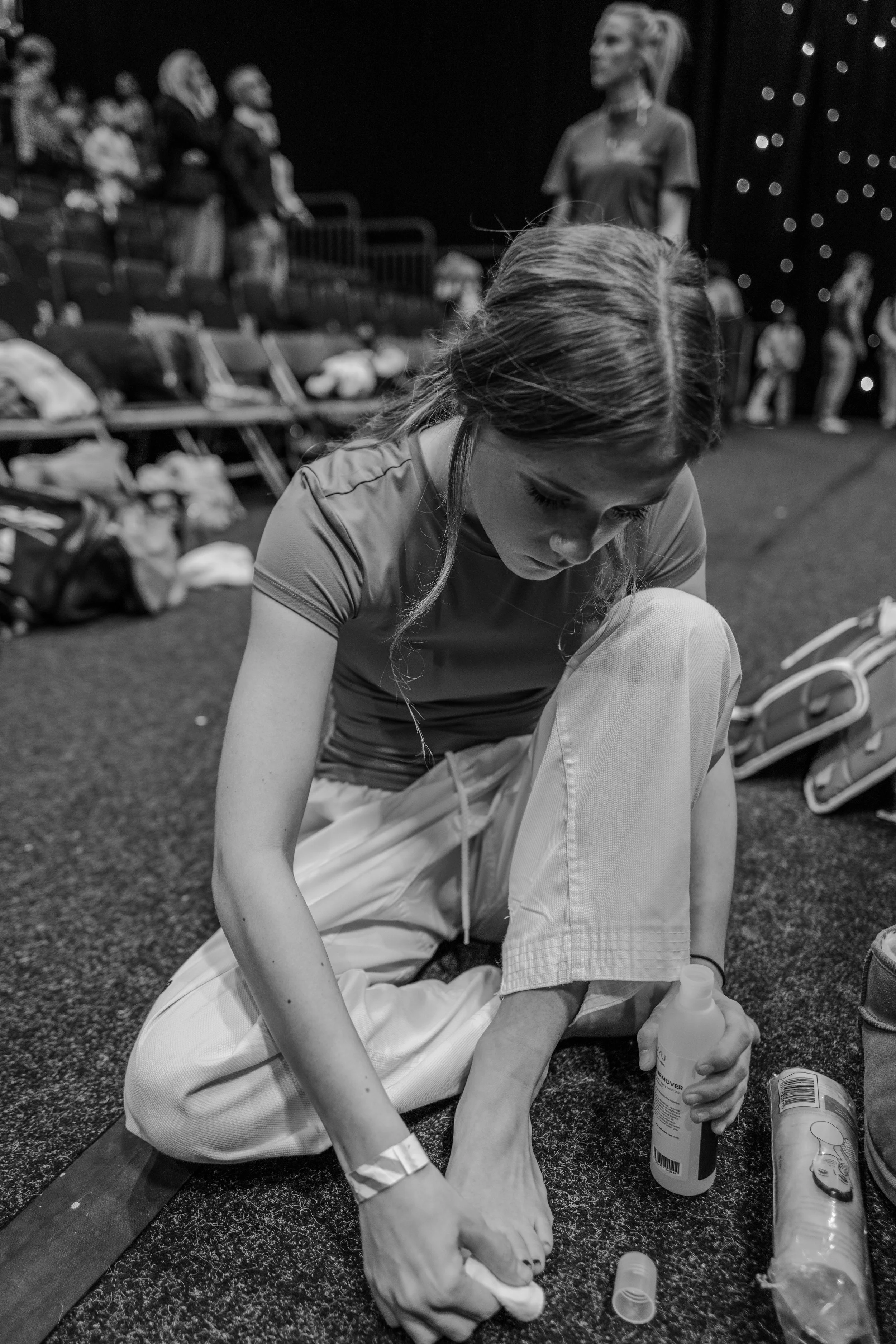A young woman with long hair tied back, sitting on the floor, cleaning her foot with a cloth and a bottle of liquid. She is surrounded by bags and other items, with an audience and people standing in the background, in what appears to be a backstage