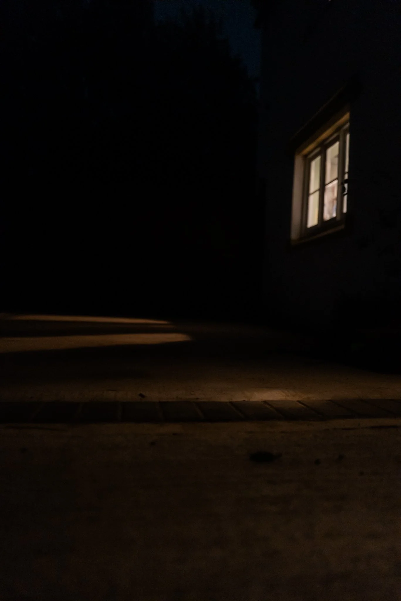 A dark outdoor scene with a single illuminated window in a building, and what appears to be a small trail or path on the ground.