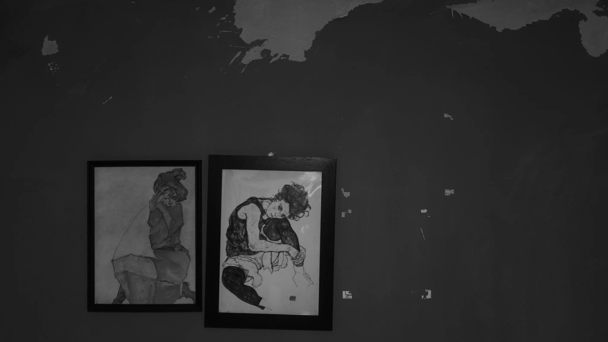 Two framed sketches of women, one with curly hair sitting with her knees up and the other with her head resting on her hand, hanging on a wall with peeling paint.
