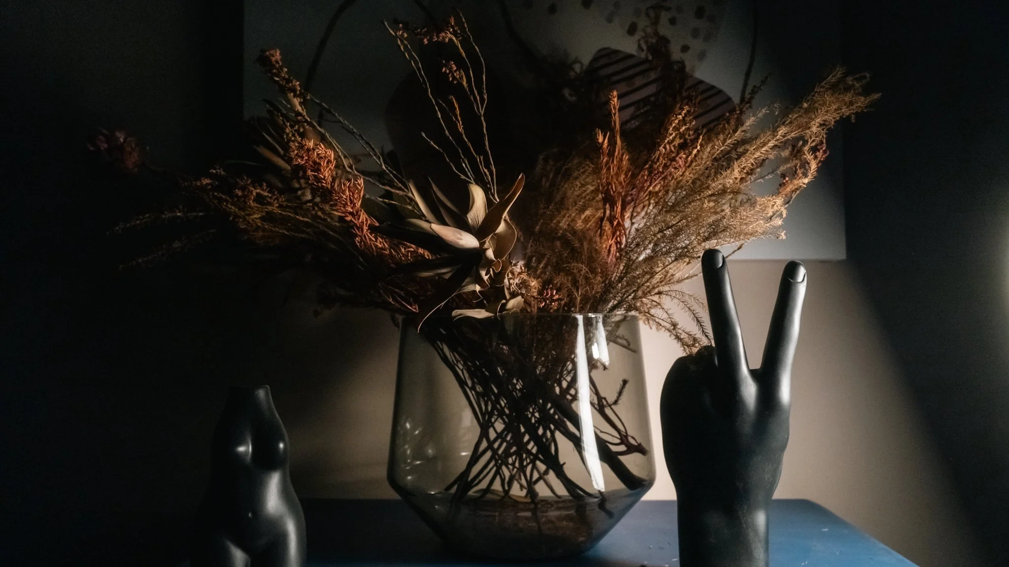 A dark-colored vase holding dried flowers and branches, with a black mannequin hand making a peace sign nearby, against a dimly lit background.