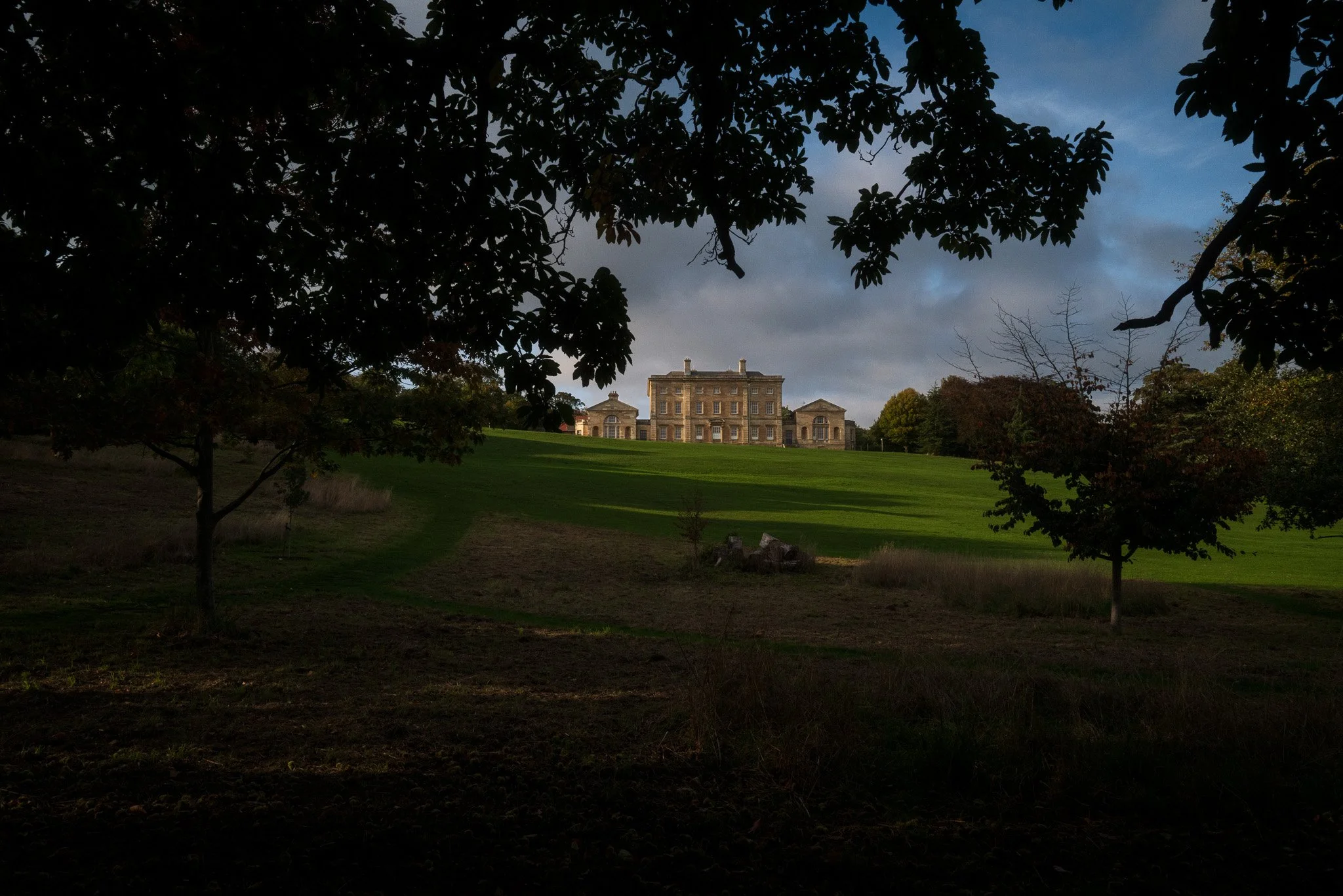 A large historic mansion on a hill, framed by trees in a park setting, under a partly cloudy sky.
