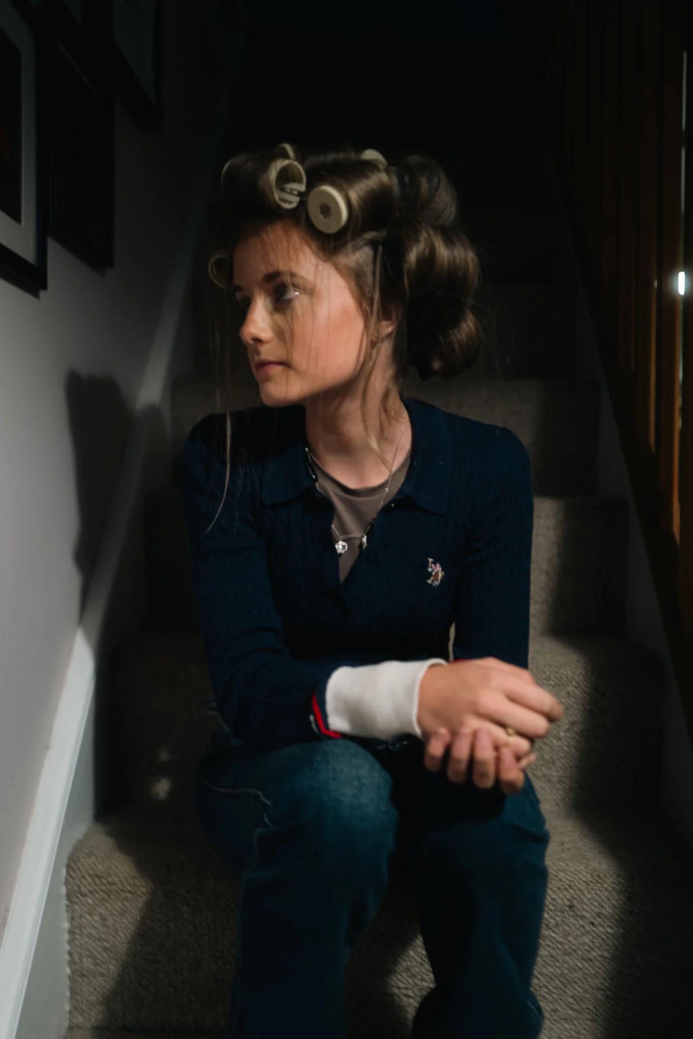 A woman sitting on a staircase with hair rollers in her hair, looking to the side, with a relaxed expression.